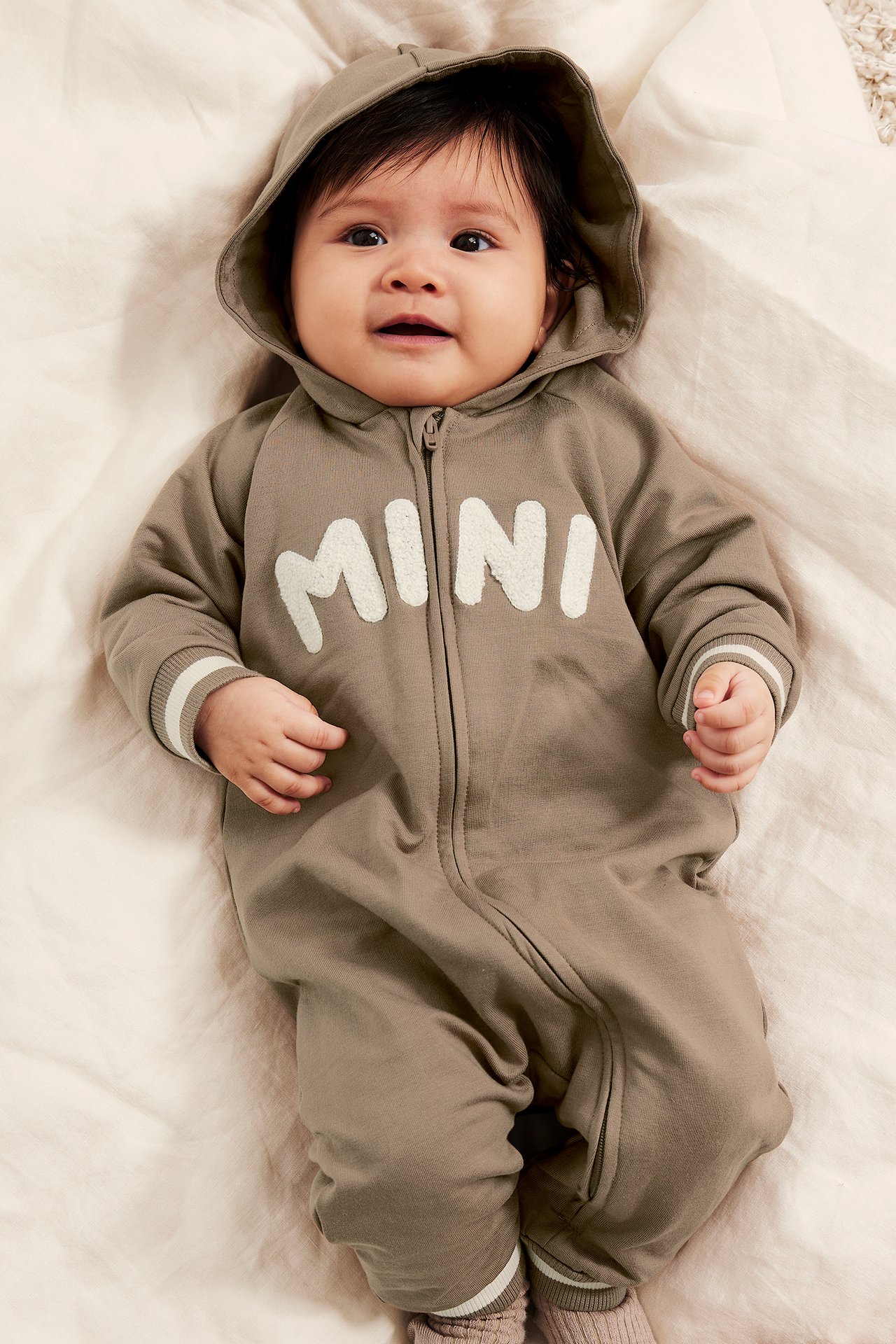 Frontvy av baby i en brun huvförsedd sweatshirtoverall med dragkedja och vitt 'MINI'-tryck.