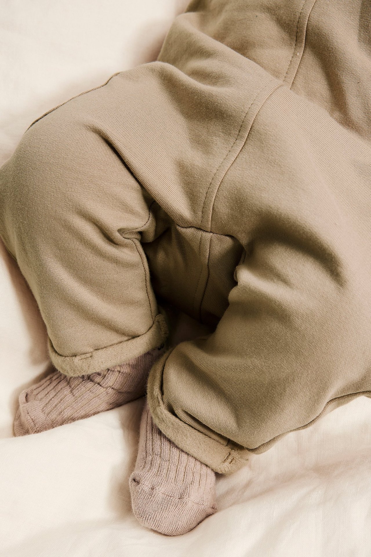 Zbliżenie na niemowlę w beżowych bawełnianych ogrodniczkach z podwiniętymi mankietami i jasnoróżowych skarpetkach w prążki.