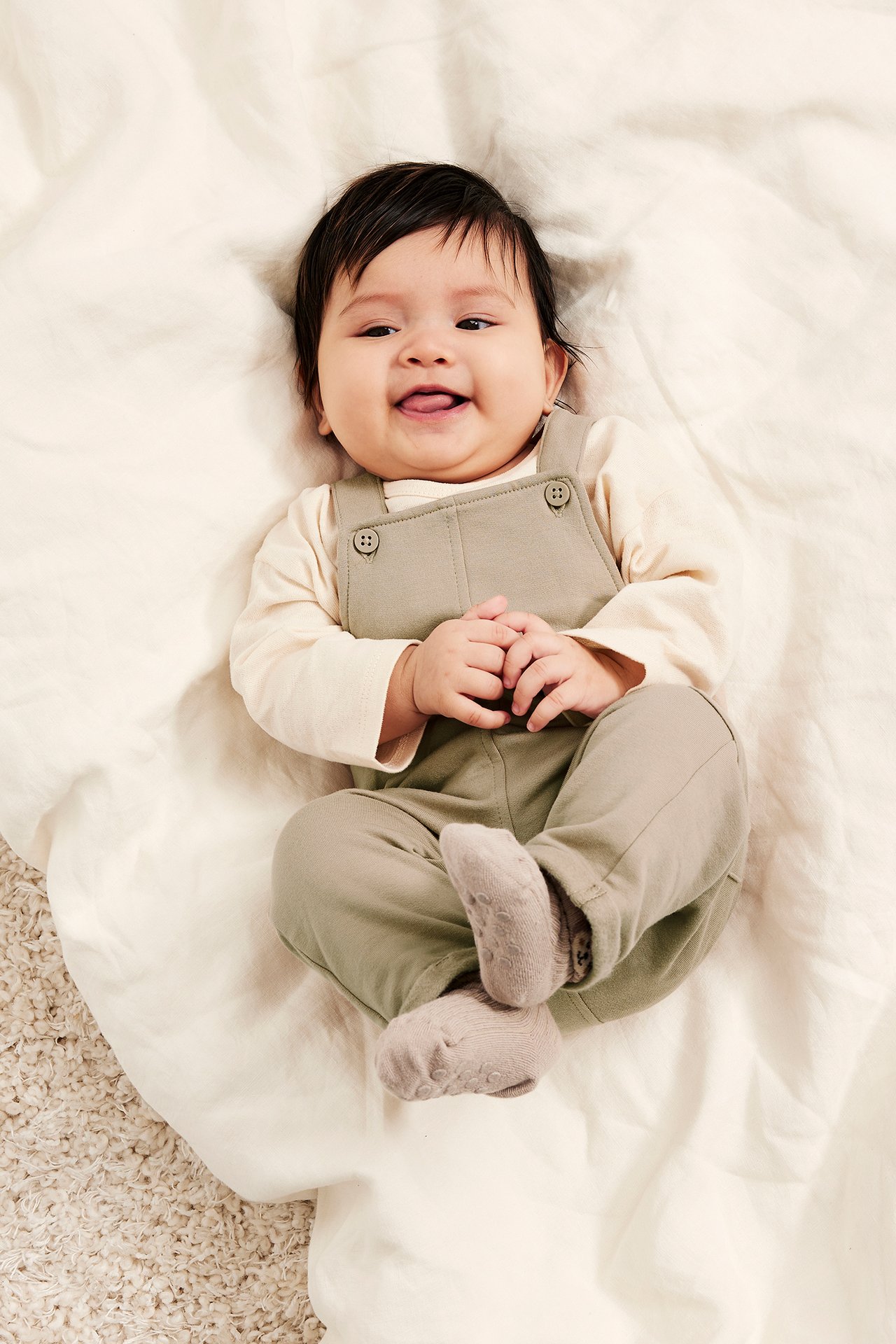 Leende baby i beige hängselbyxor med knappar över en krämfärgad långärmad topp och ljusgrå strumpor.