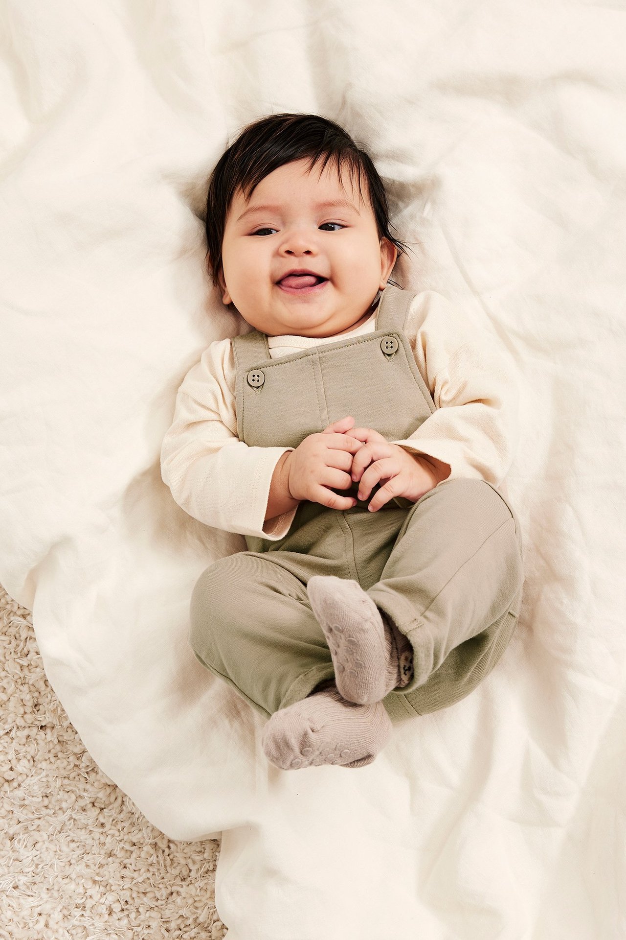 Uśmiechnięte dziecko w beżowych ogrodniczkach z guzikami, na kremowej bluzce i jasnoszarych skarpetkach.