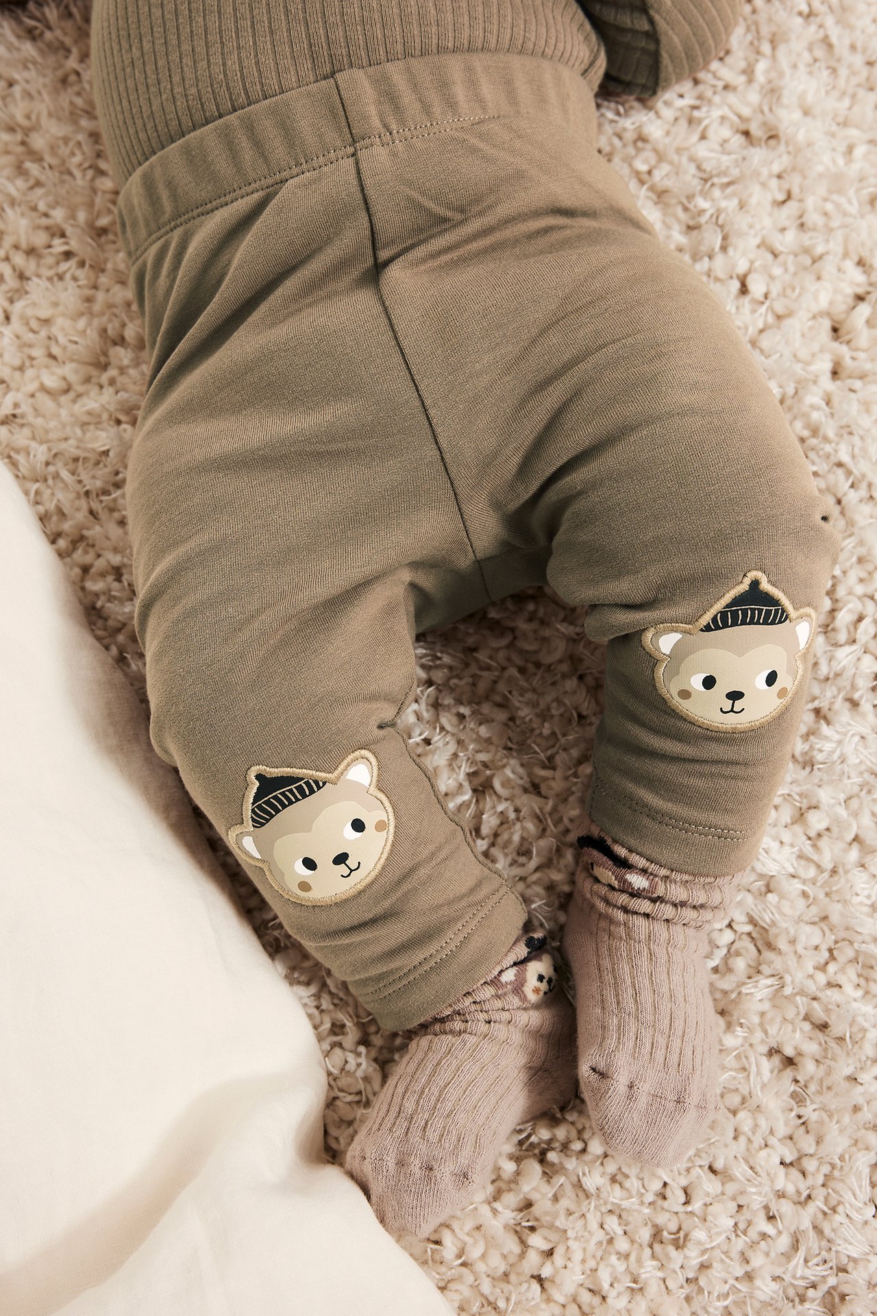 Lähikuva vauvasta ruskeissa leggingseissä, joissa karhusovellukset polvissa, ribattu ruskea toppi ja vaaleanruskeat sukat.