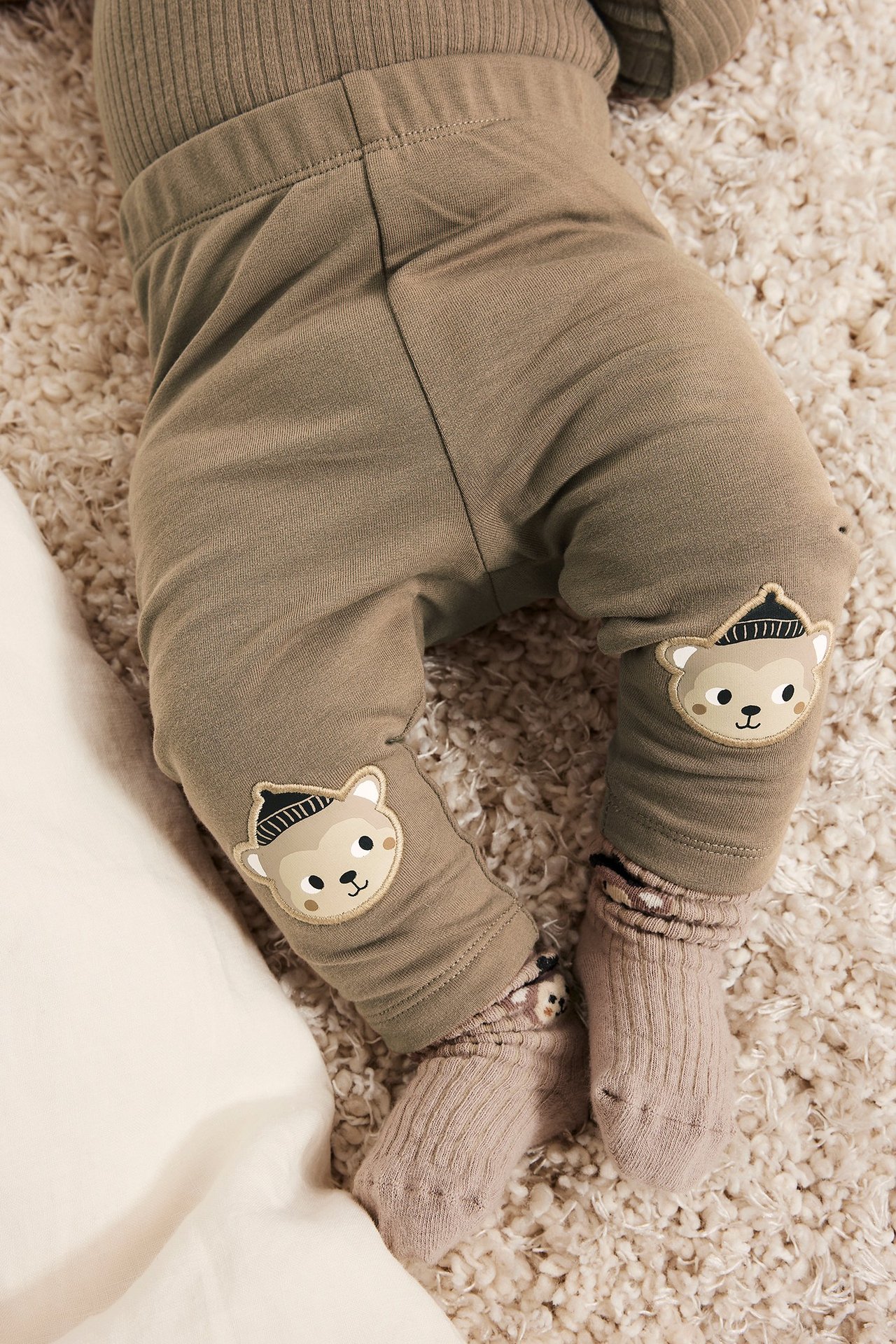 Lähikuva vauvasta ruskeissa leggingseissä, joissa karhusovellukset polvissa, ribattu ruskea toppi ja vaaleanruskeat sukat.