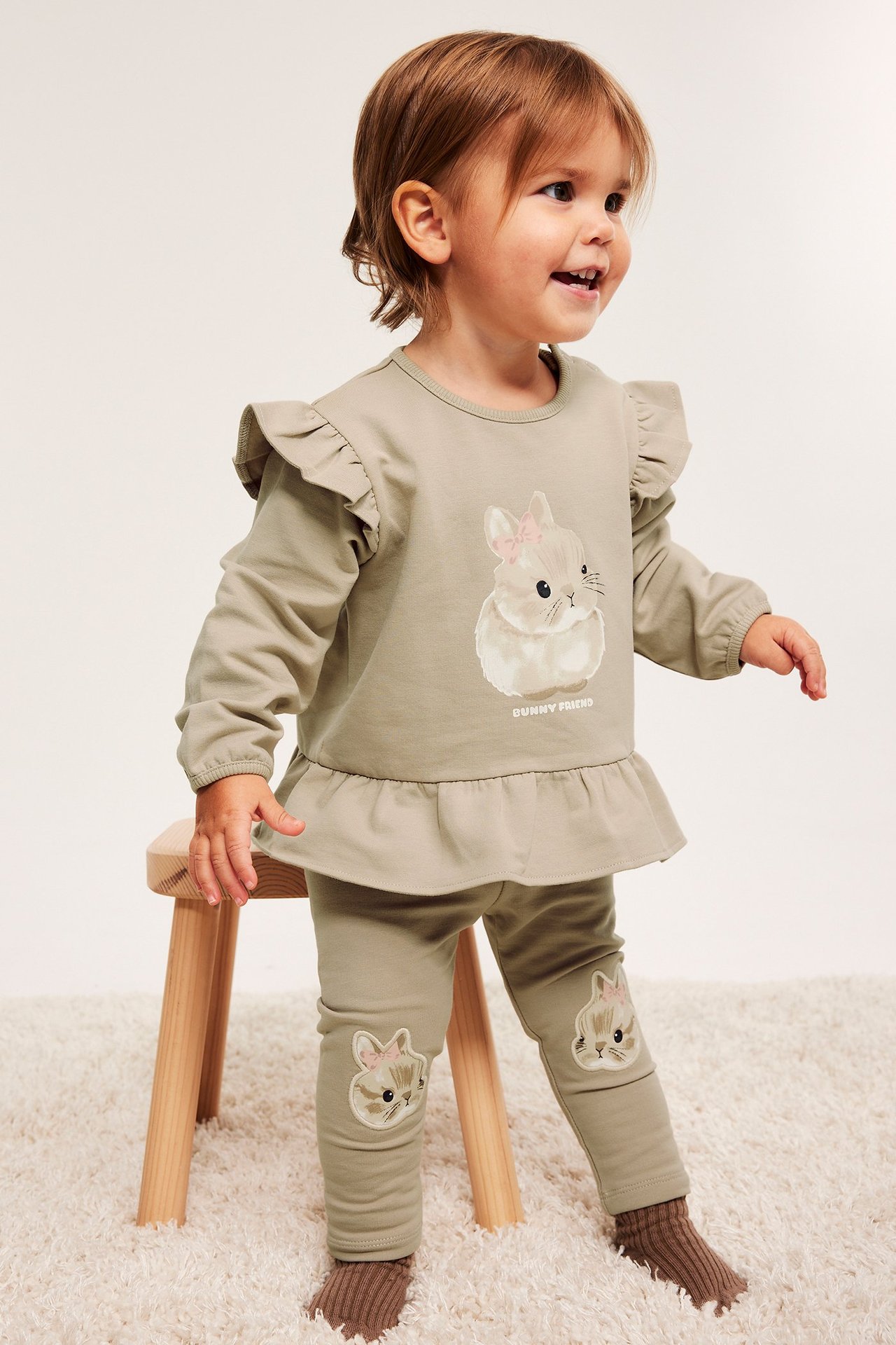Frontvy: Baby i beige leggings med kaninapplikationer, beige volangtopp med kanintryck, och bruna strumpor.