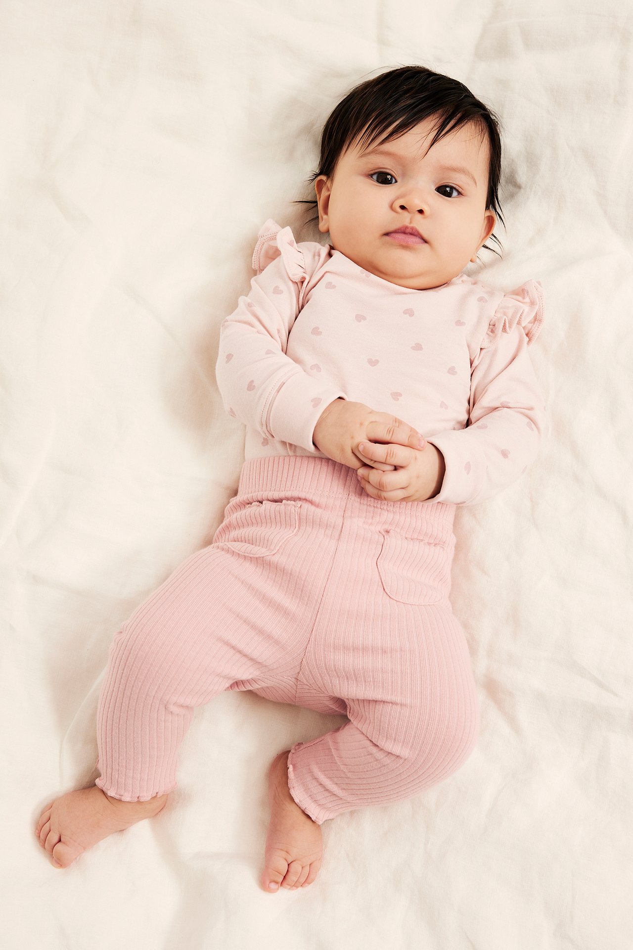 Framifrån, baby i rosa ribbade leggings och en rosa långärmad body med hjärtmönster.