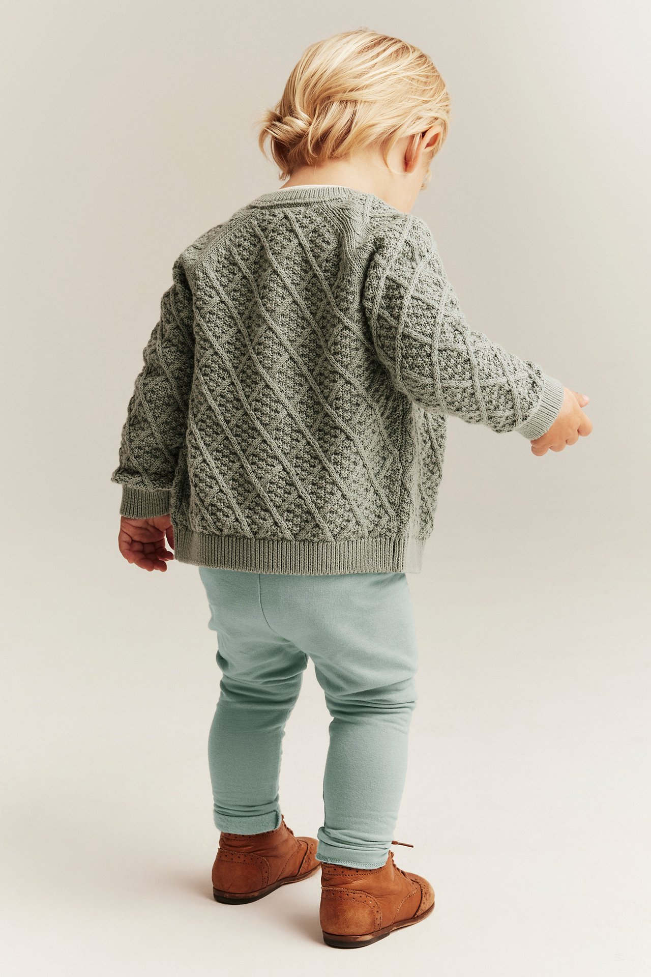 Ryggvisning av baby i lysegrønne leggings, en grå diamantmønstret strikket cardigan og brune snørestøvler.