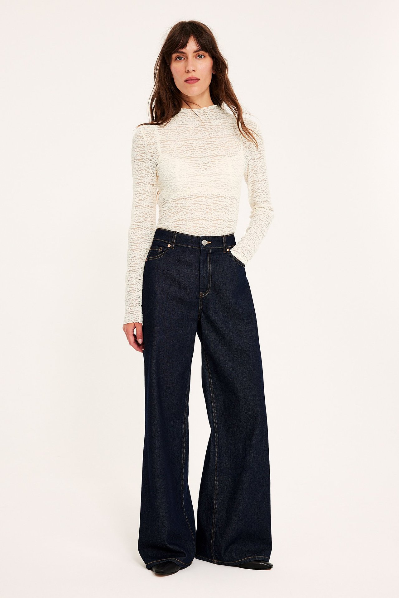 Edestä: nainen yllään luonnonvalkoinen pitsinen pitkähihainen toppi ja tummansiniset leveälahkeiset farkut.