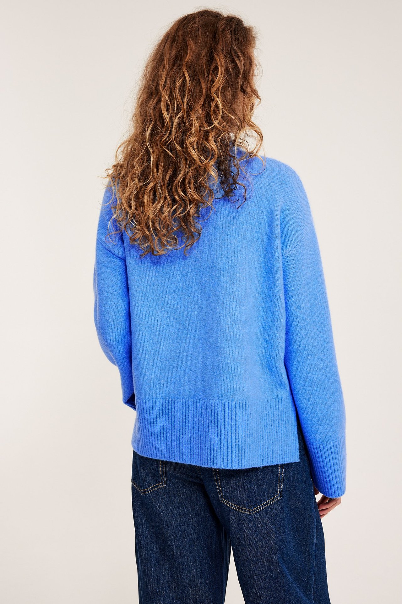 Baksidan av en kvinna som bär en blå stickad pullover med ribbad nederkant och mörkblå jeans.