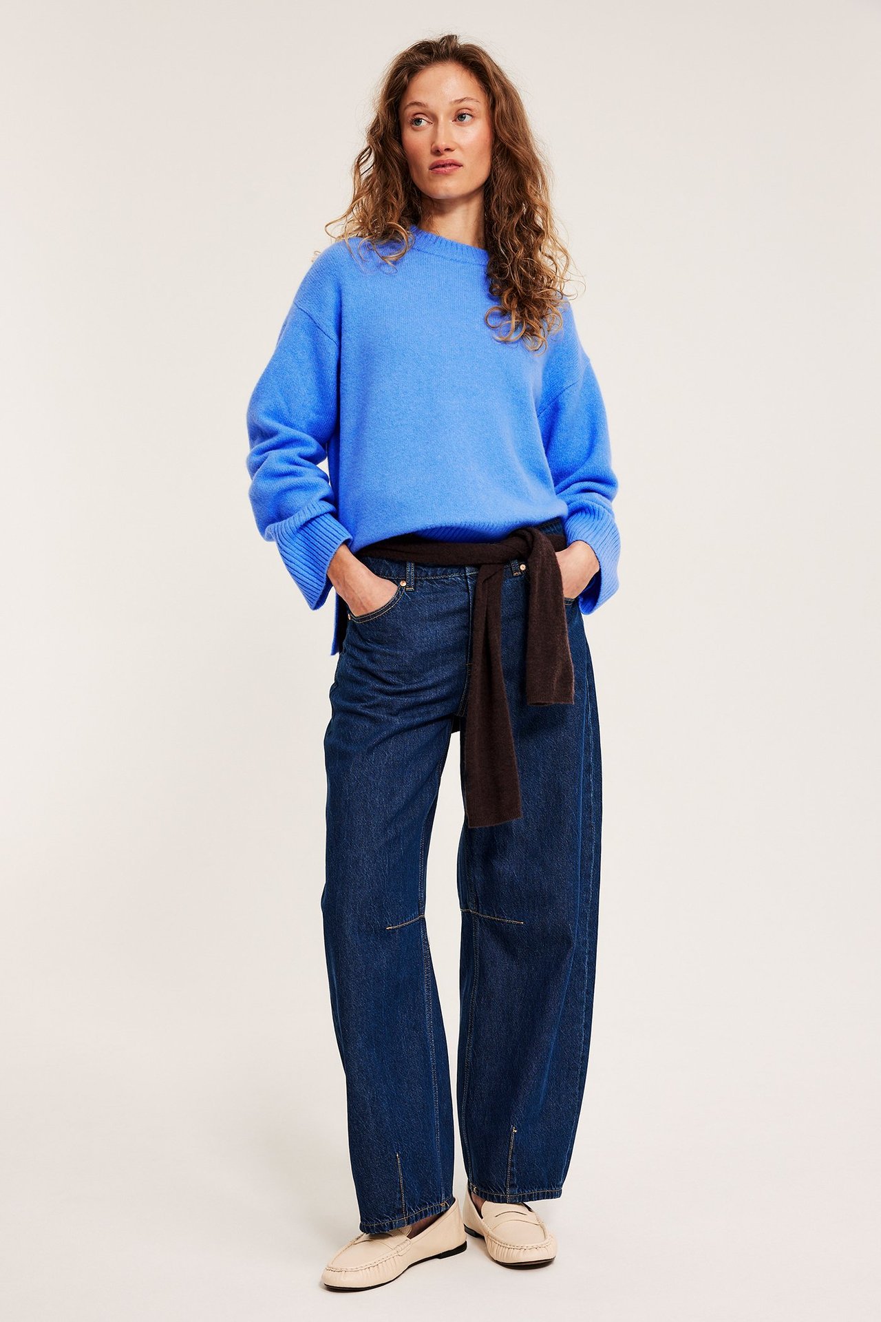 Frontalvisning: Kvinne i blå strikket genser, mørkeblå jeans, beige loafers; brun genser knyttet rundt livet.