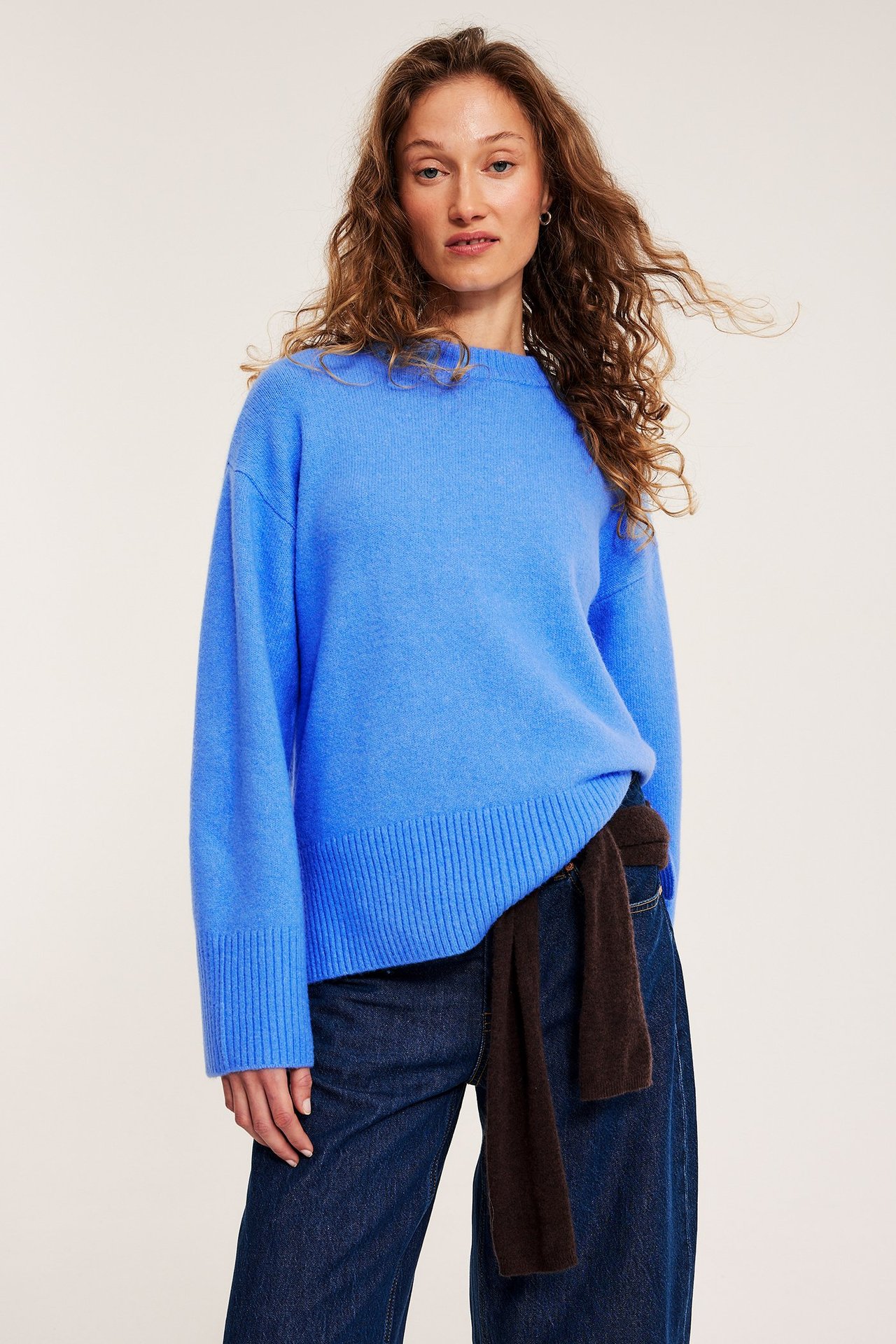 Frontvisning: Kvinne i blå strikket rundhalset genser, mørke jeans og brunt plagg knyttet i midjen.