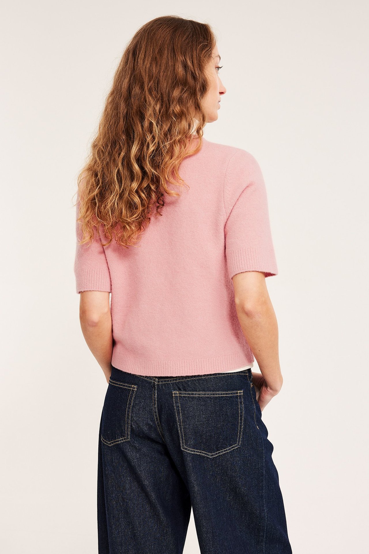 Nainen selin, yllään vaaleanpunainen lyhythihainen neulepusero ja tummansiniset farkut.