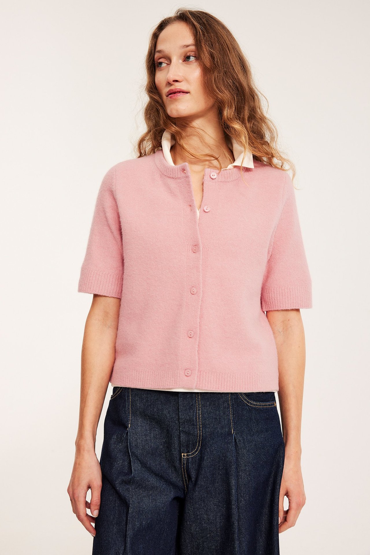 En kvinne i rosa kortermet strikket cardigan, hvit topp og mørkeblå vide jeans, front.