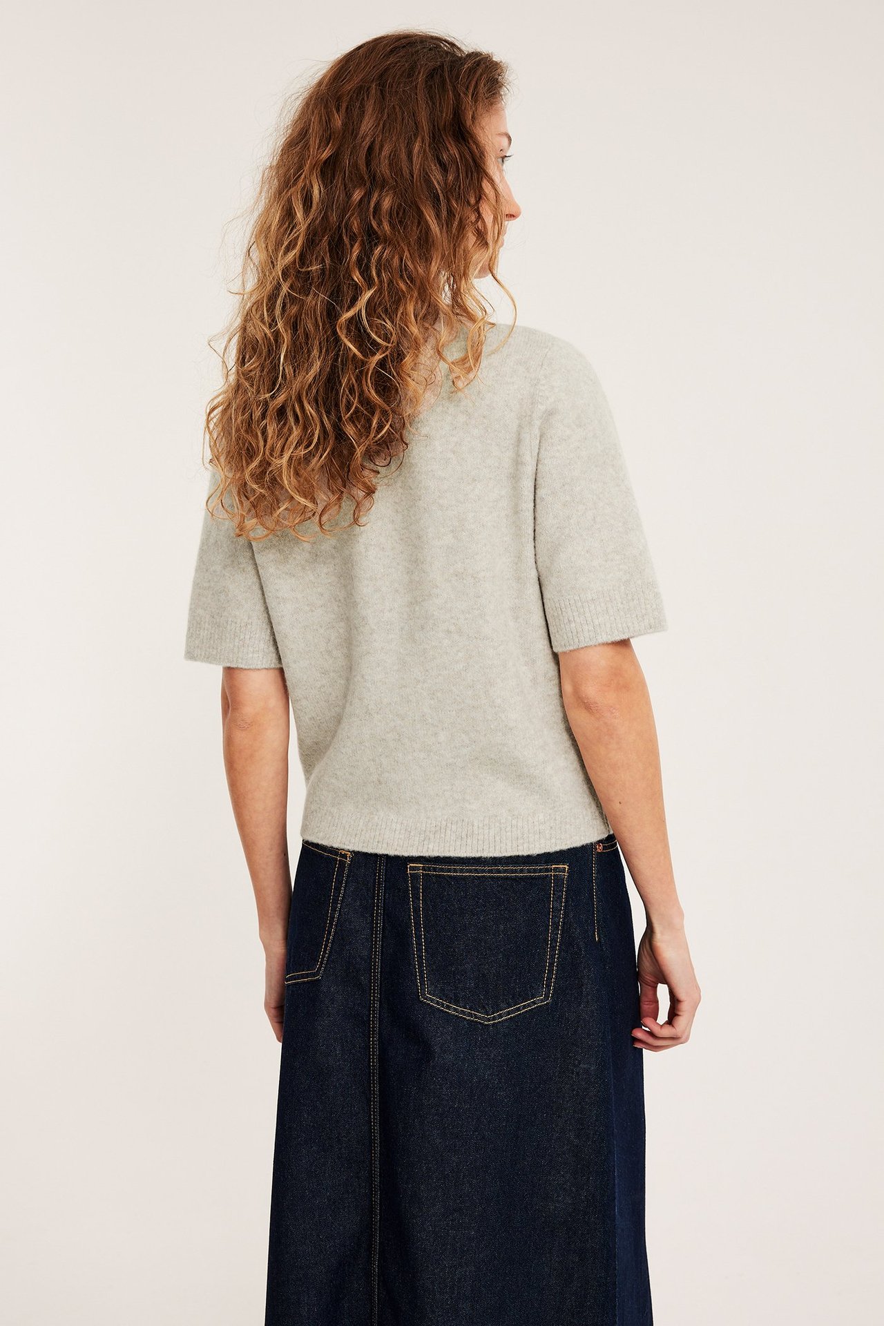 Naisen takakuva yllään vaaleanbeige meleerattu lyhythihainen neulepusero ja tummansininen farkkuhame.