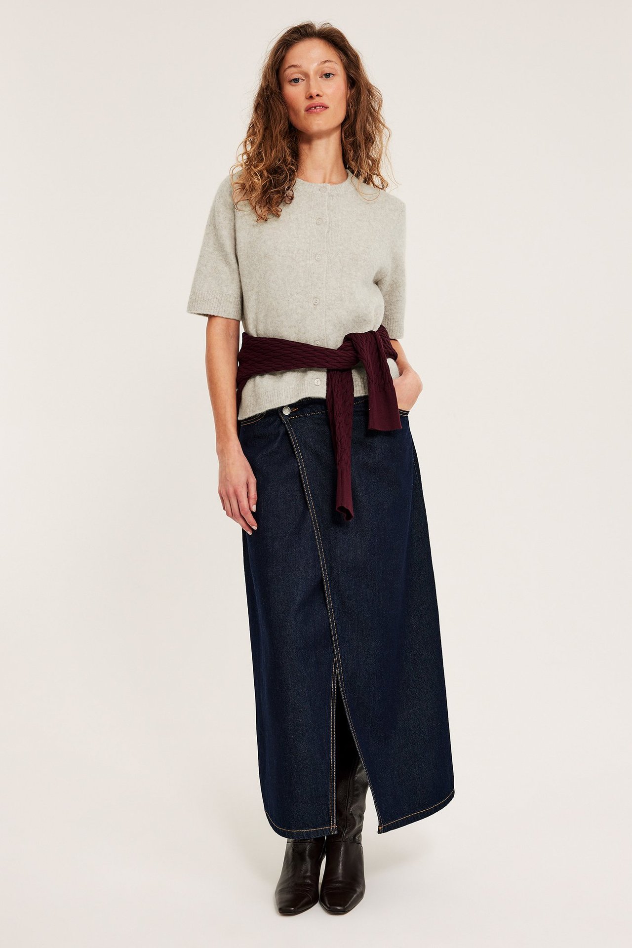 Edestä: Nainen vaaleanbeigessä meleeratussa lyhythihaisessa neuletakissa, tummansinisessä farkkuhameessa ja tummissa saappaissa. Viininpunainen neule vyötäröllä.