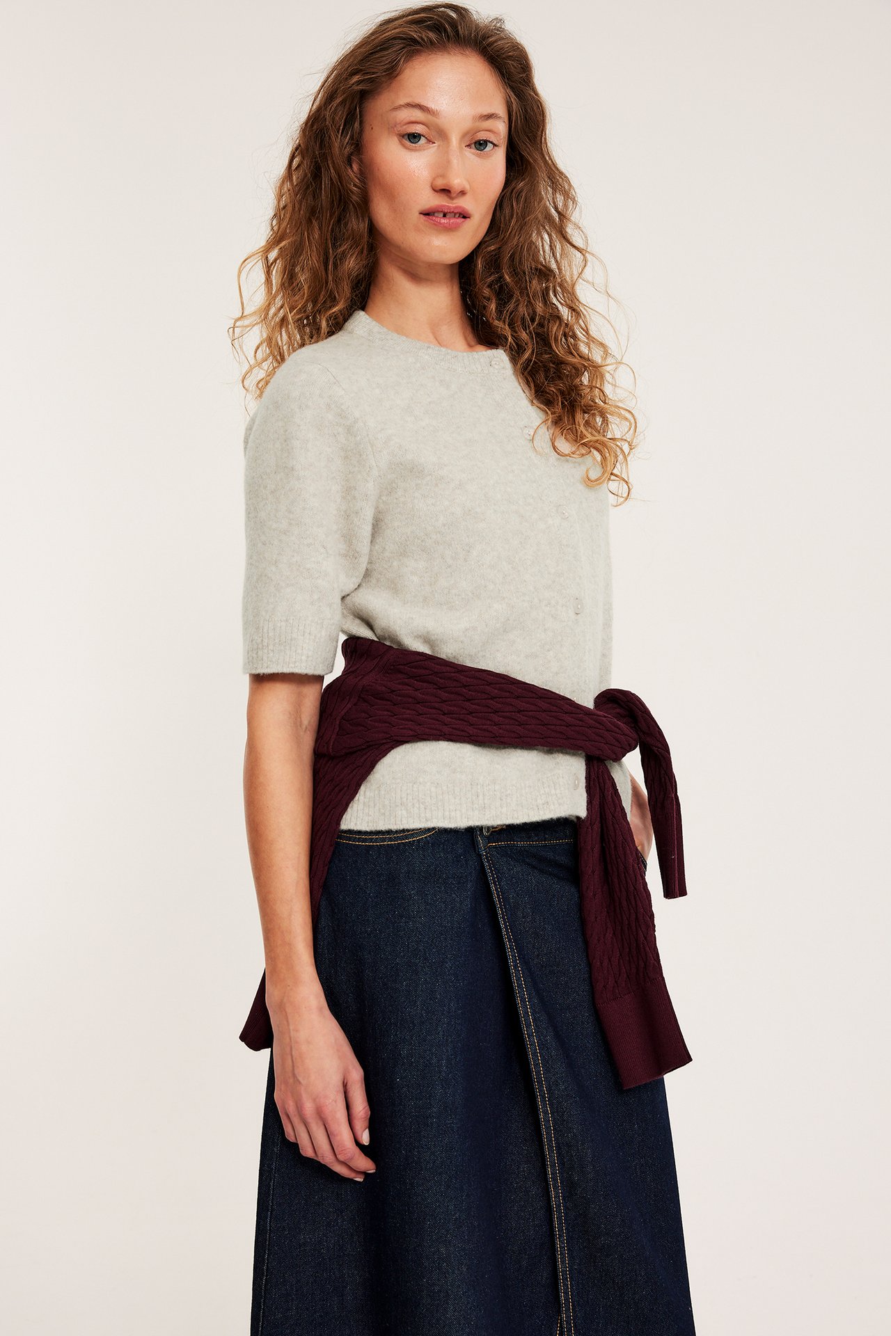 Nainen melange vaaleanbeigessä lyhythihaisessa neuletakissa, tummassa farkkuhameessa ja luumunvärisessä neuleessa vyötärölle sidottuna.