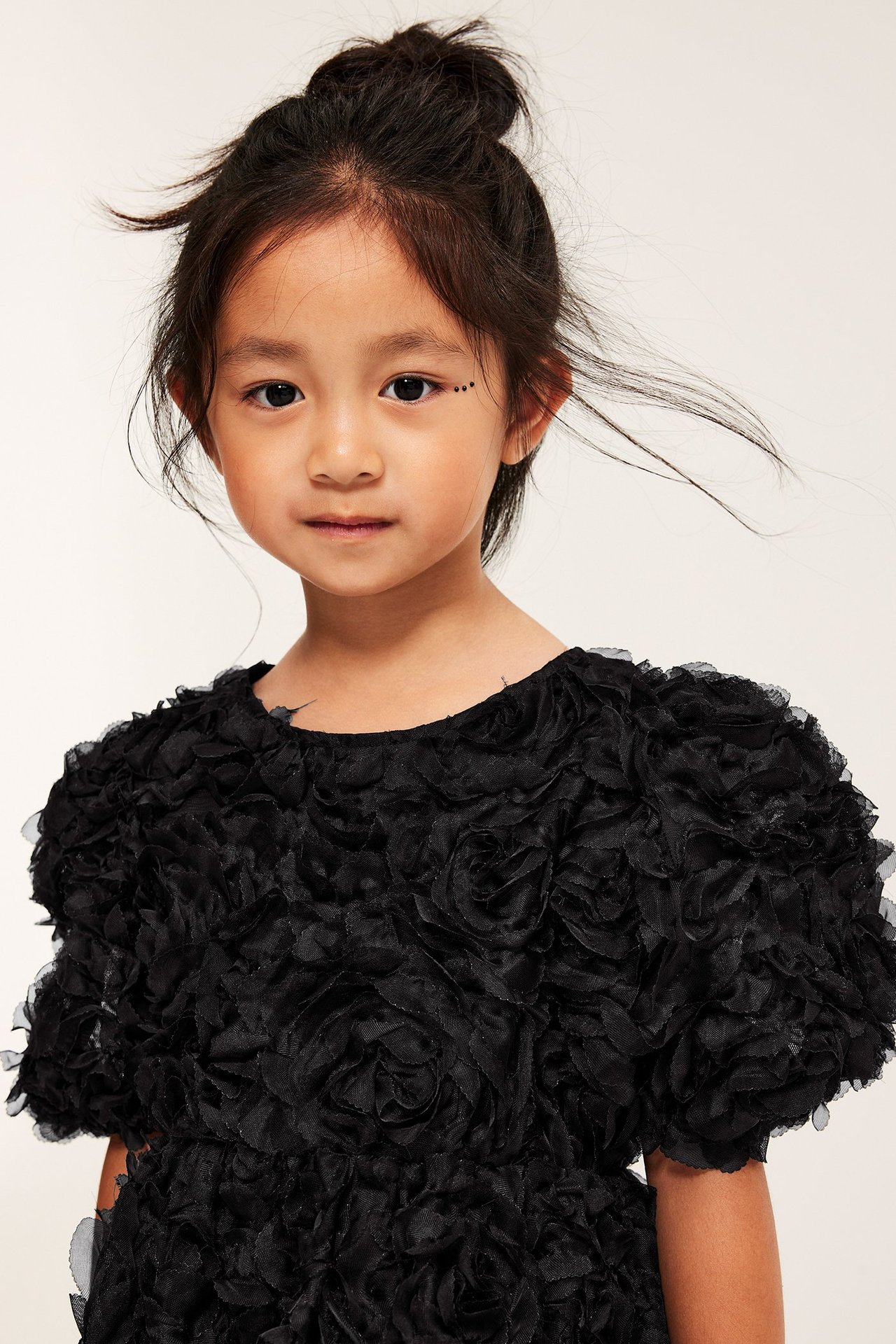 Lähikuva, edestä, lapsi yllään musta mekko teksturoituilla 3D-ruusuilla ja pöyhkeillä hihoilla.