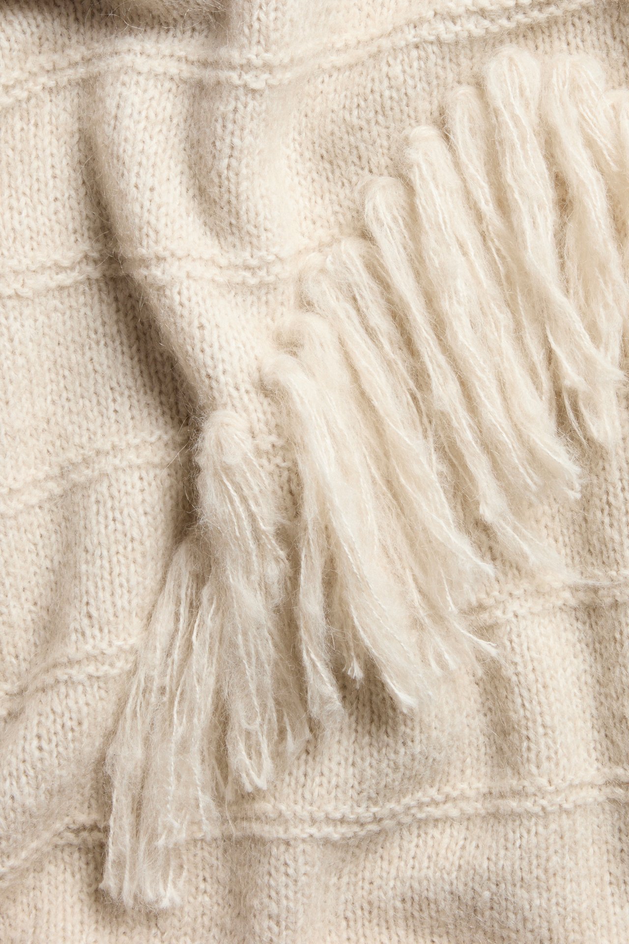 Nærbilde av en lys beige grovstrikket genser i ullblanding med fluffy frynsedetaljer.