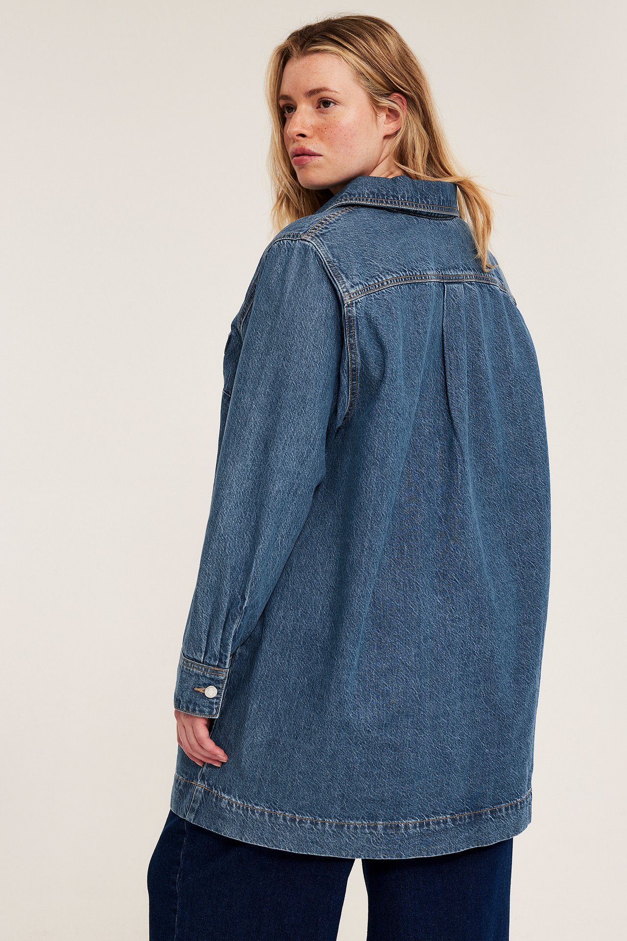 Widok z tyłu kobiety w luźnej niebieskiej sukience koszulowej z jeansu na ciemnoniebieskich spodniach.