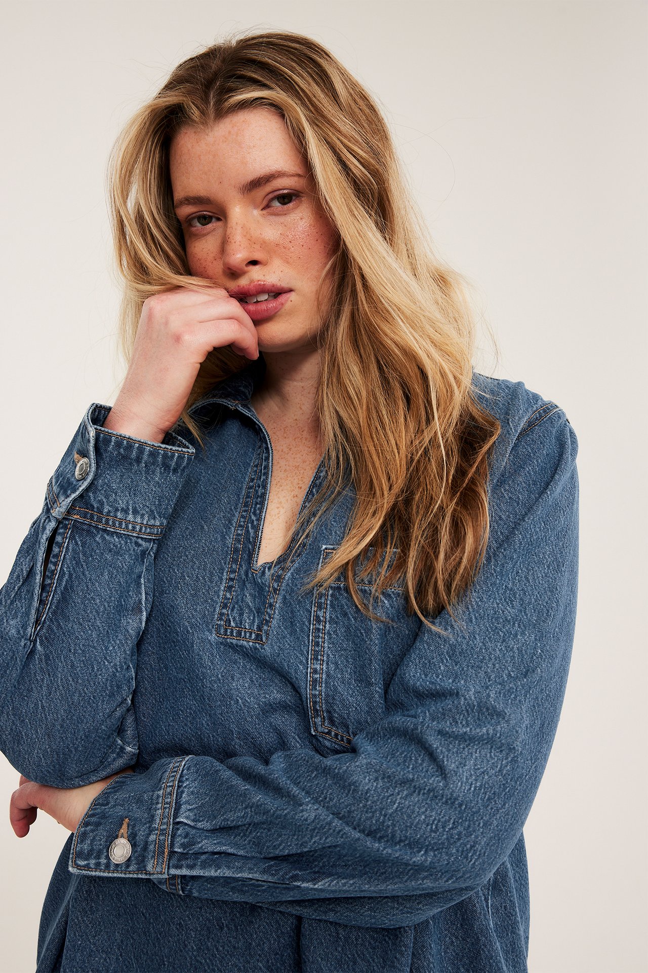 Lähikuva edestä naisesta, jolla on väljä sininen farkkumekko V-pääntiellä ja napitetuilla hihansuilla.