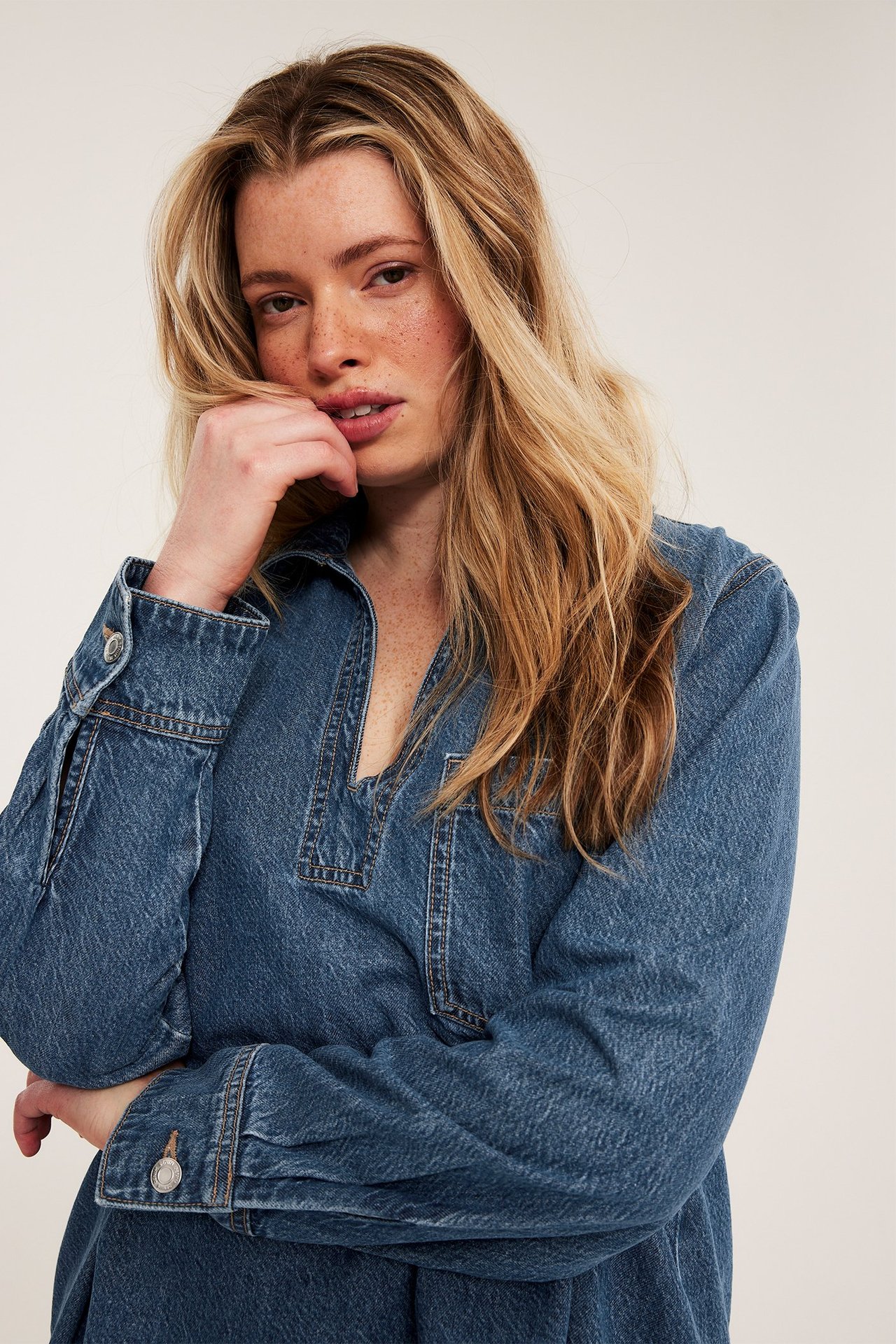 Närbild framifrån på kvinna i lös blå jeansklänning med V-ringning och knäppta manschetter.