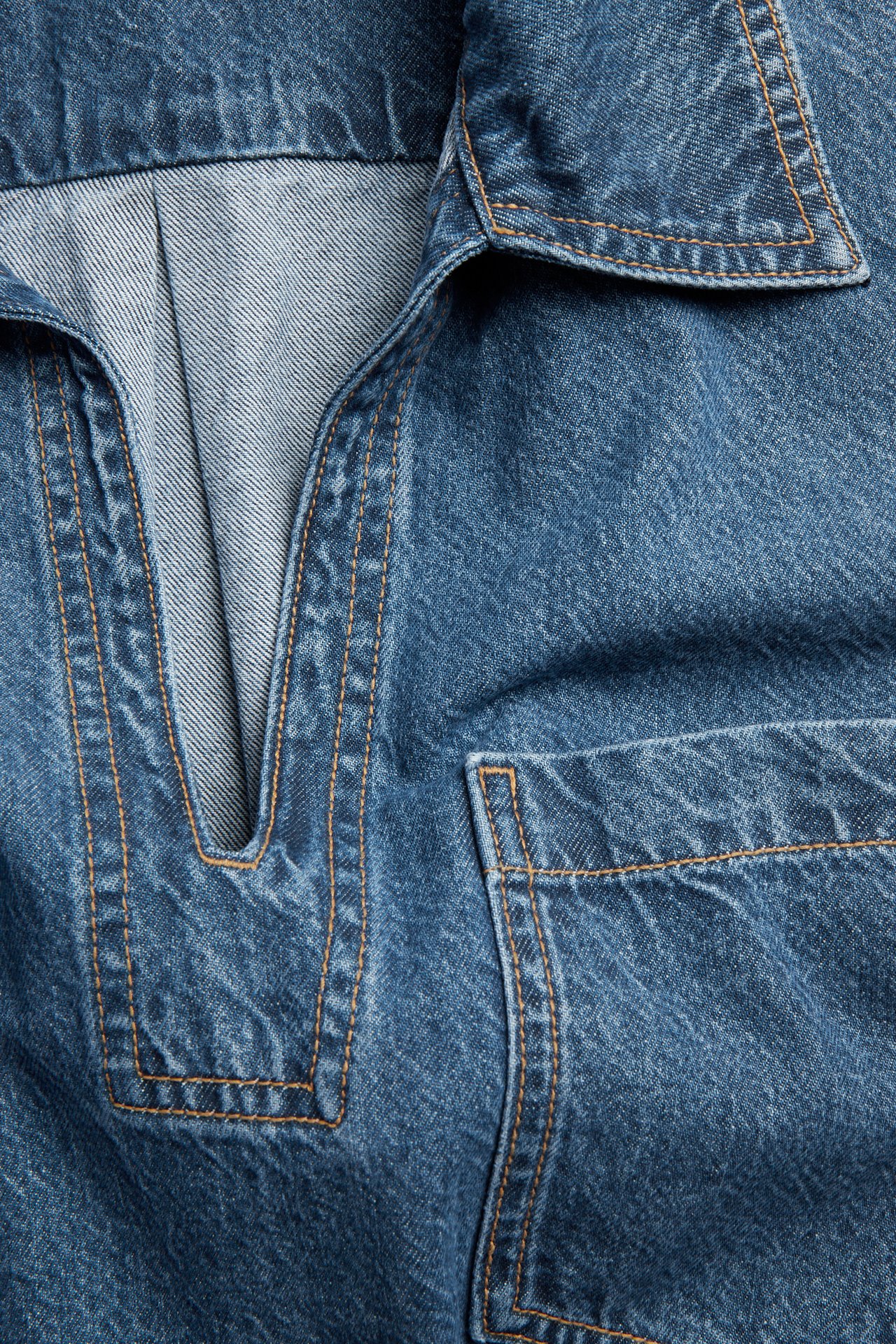 Närbild av en blå jeansklänning med V-ringning, krage, bröstficka och orange sömmar.