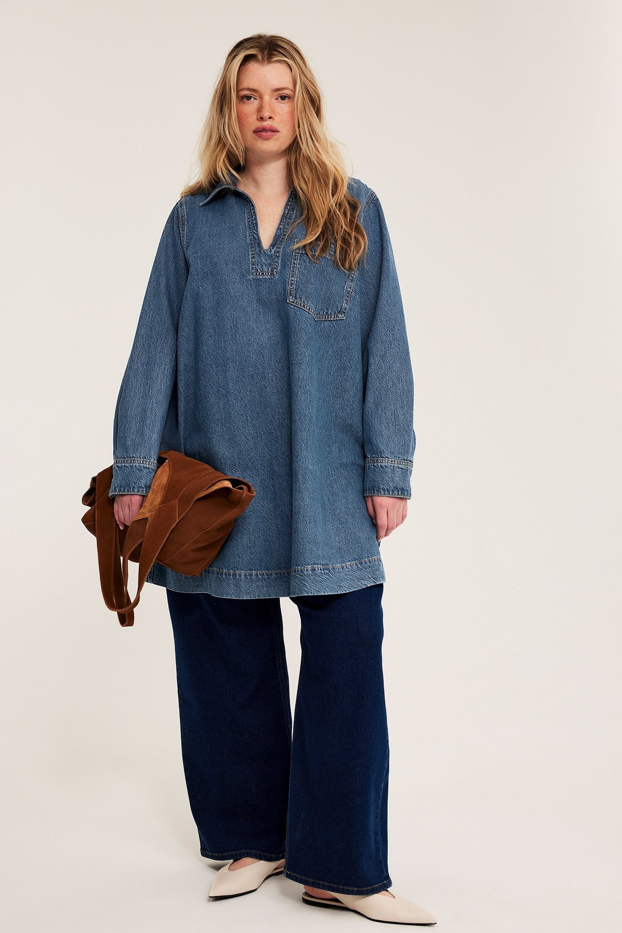 Framifrån: Kvinna i ljusblå denimtunika, mörkblå vida jeans, offwhite-tofflor, håller brun väska.