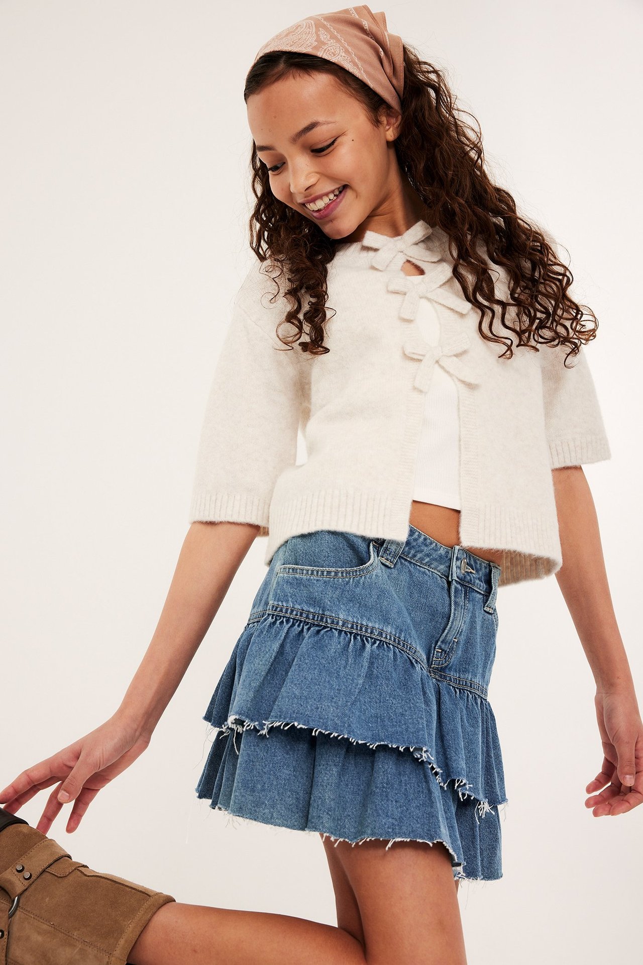 Barn bär en ljusblå volangjeanskjol, en vit rosettkofta och en mönstrad beige bandana.