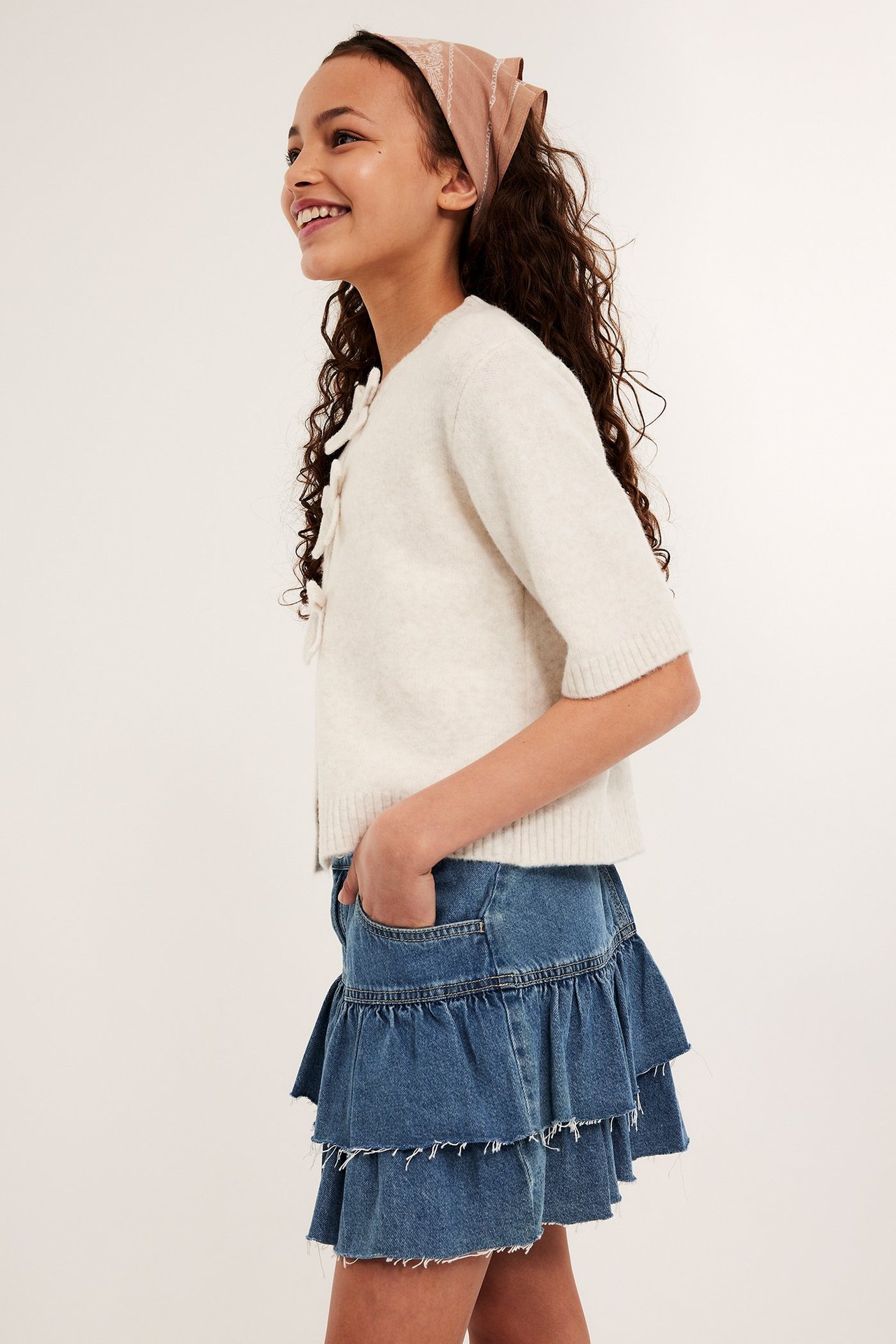 Widok z boku uśmiechniętego dziecka w jasnoniebieskiej jeansowej spódniczce, beżowym swetrze i brązowej bandanie.