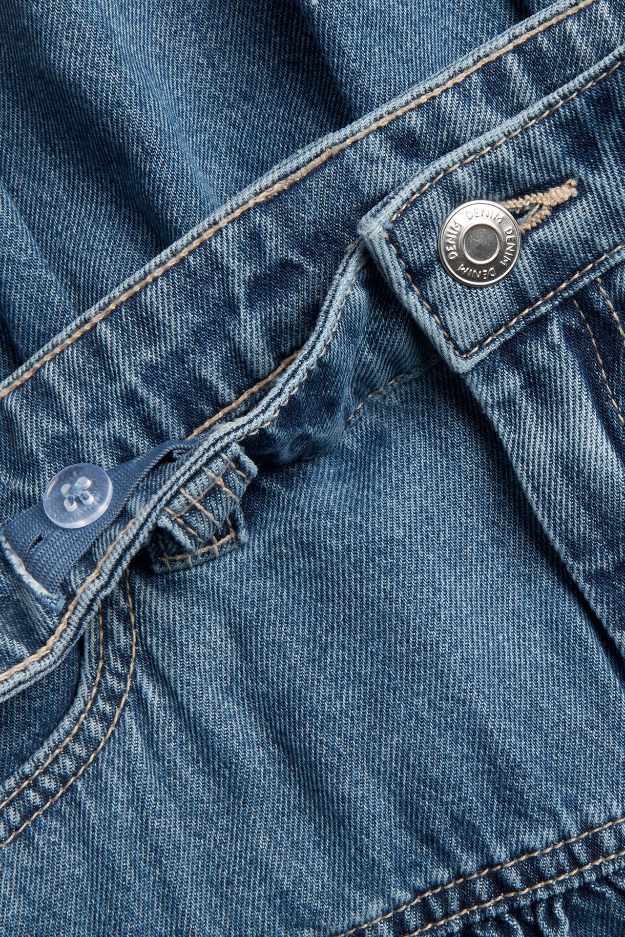 Nærbilde av et lys blått denimskjørt for barn med rysjer, metallknapp, glidelås og justerbar midje.