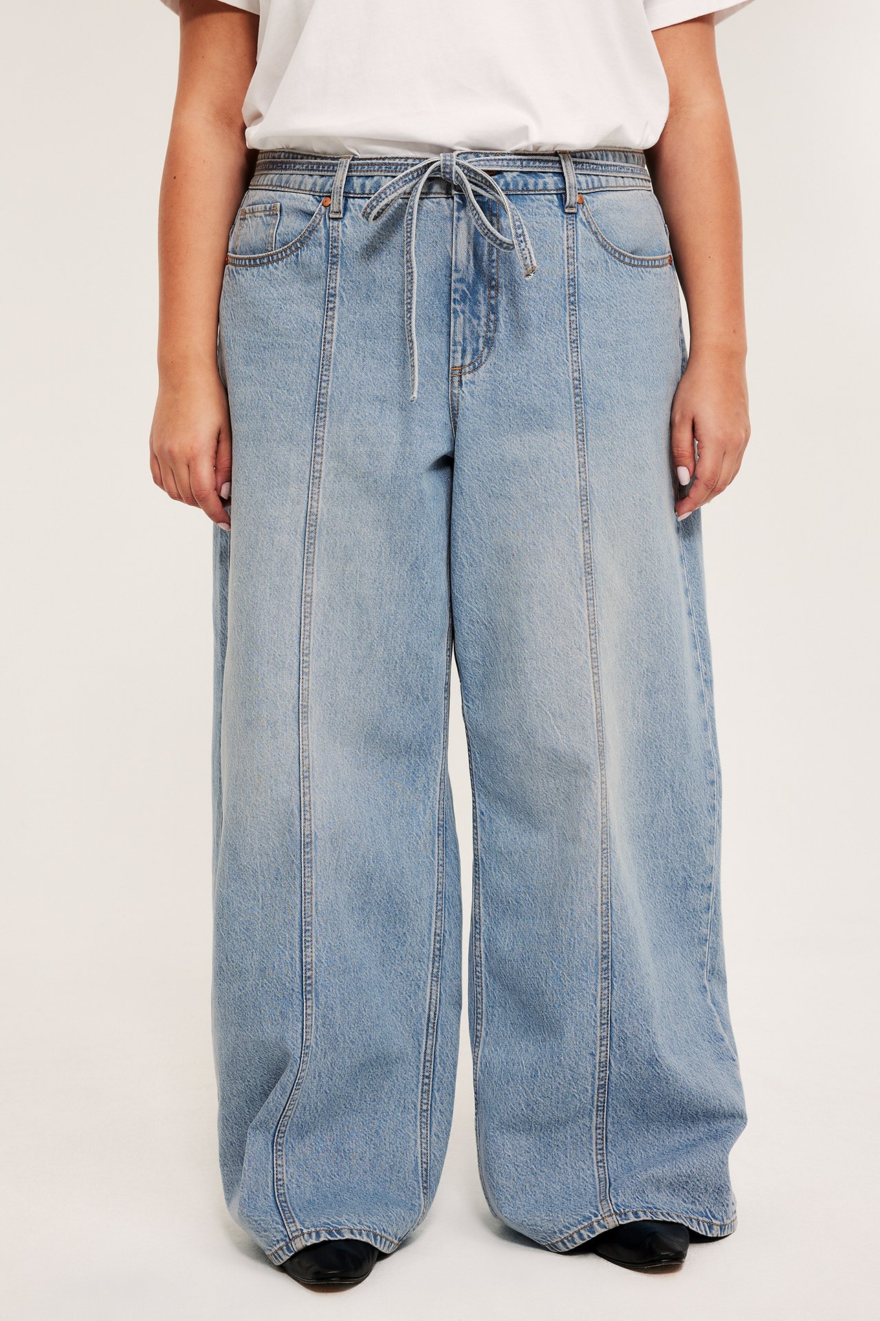 Framifrån: Kvinna i ljusblå vida jeans med hög midja, framsömmar och dragsko, matchad med vit t-shirt och svarta skor.