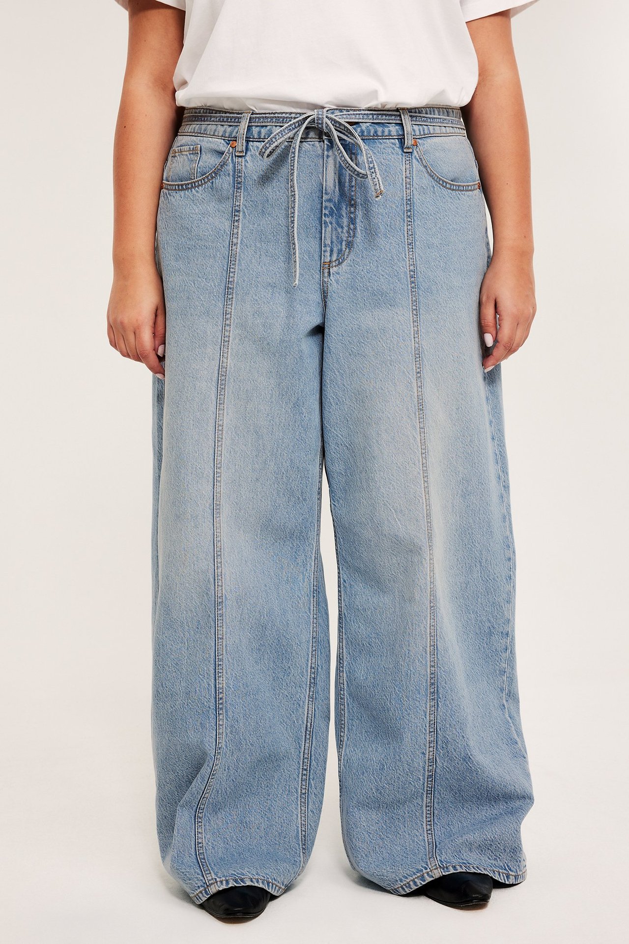 Etunäkymä: Nainen yllään vaaleansiniset leveälahkeiset korkeavyötäröiset farkut etusaumoilla ja kiristysnyörillä, valkoinen t-paita ja mustat kengät.