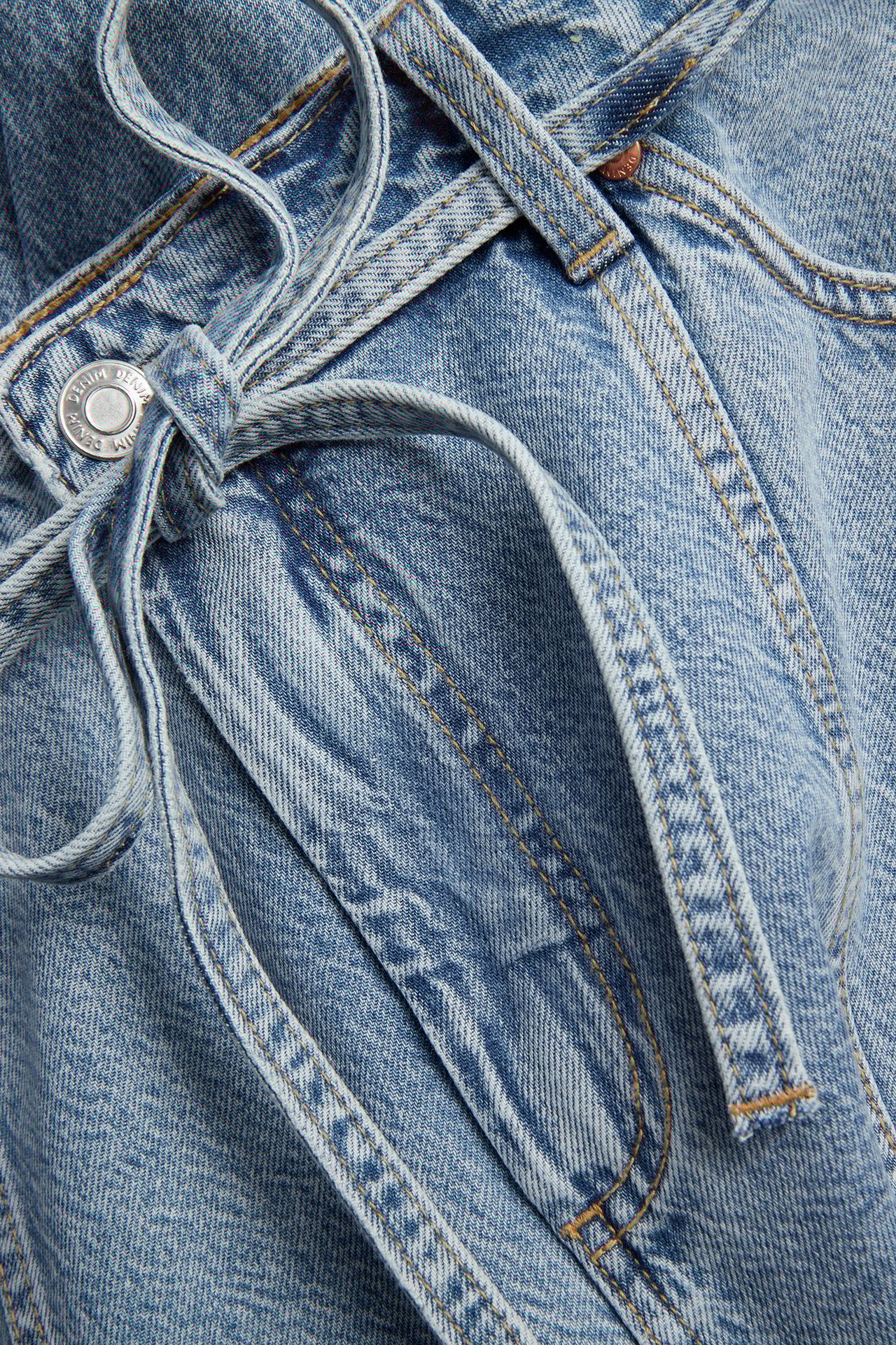 Närbild av ljusblå vida denimjeans med sömmar framtill och ett matchande denimskärp knutet i en rosett.