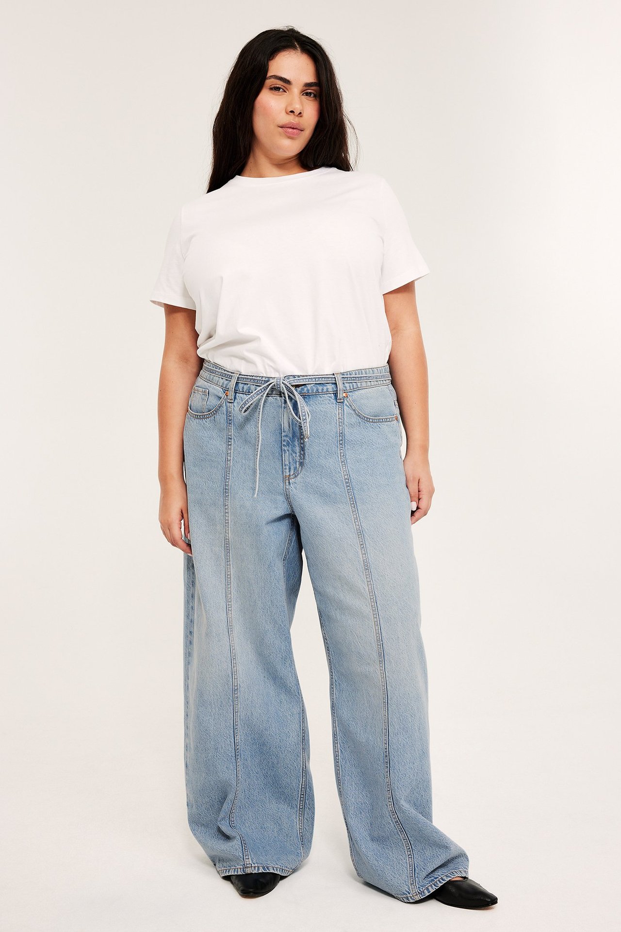 Frontvy: Kvinna i ljusblå vida jeans med sömmar, dragsko i midjan, vit t-shirt och svarta skor.