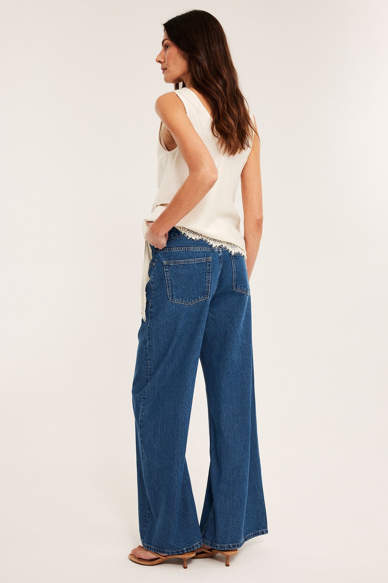 Sivukuva naisesta sinisissä leveissä korkeavyötäröisissä farkuissa, luonnonvalkoisessa topissa ja ruskeissa korkosandaaleissa.