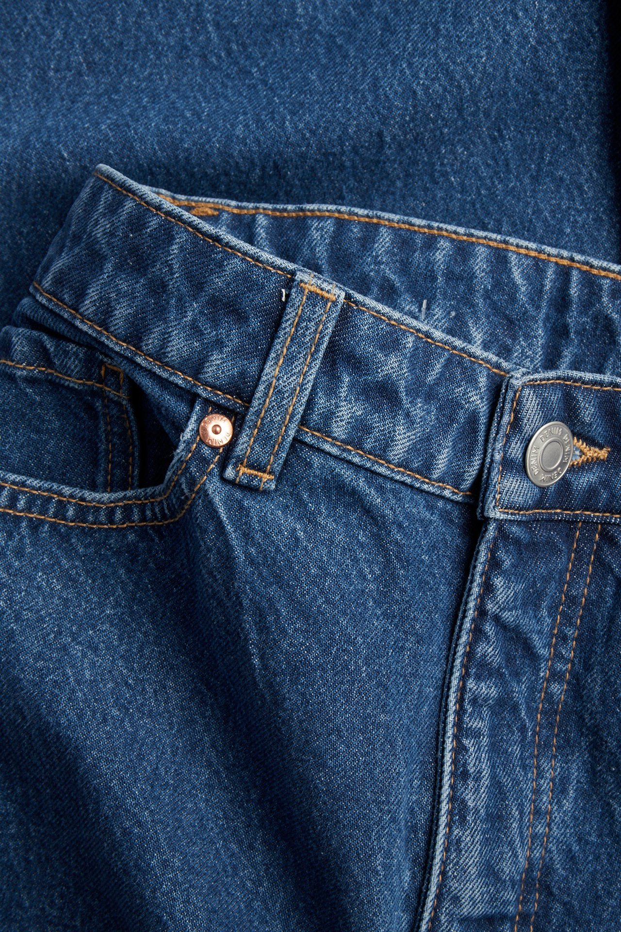 Lähikuva edestä sinisistä denimin korkeavyötäröisistä leveistä farkuista oransseilla tikkauksilla ja metallinapilla.