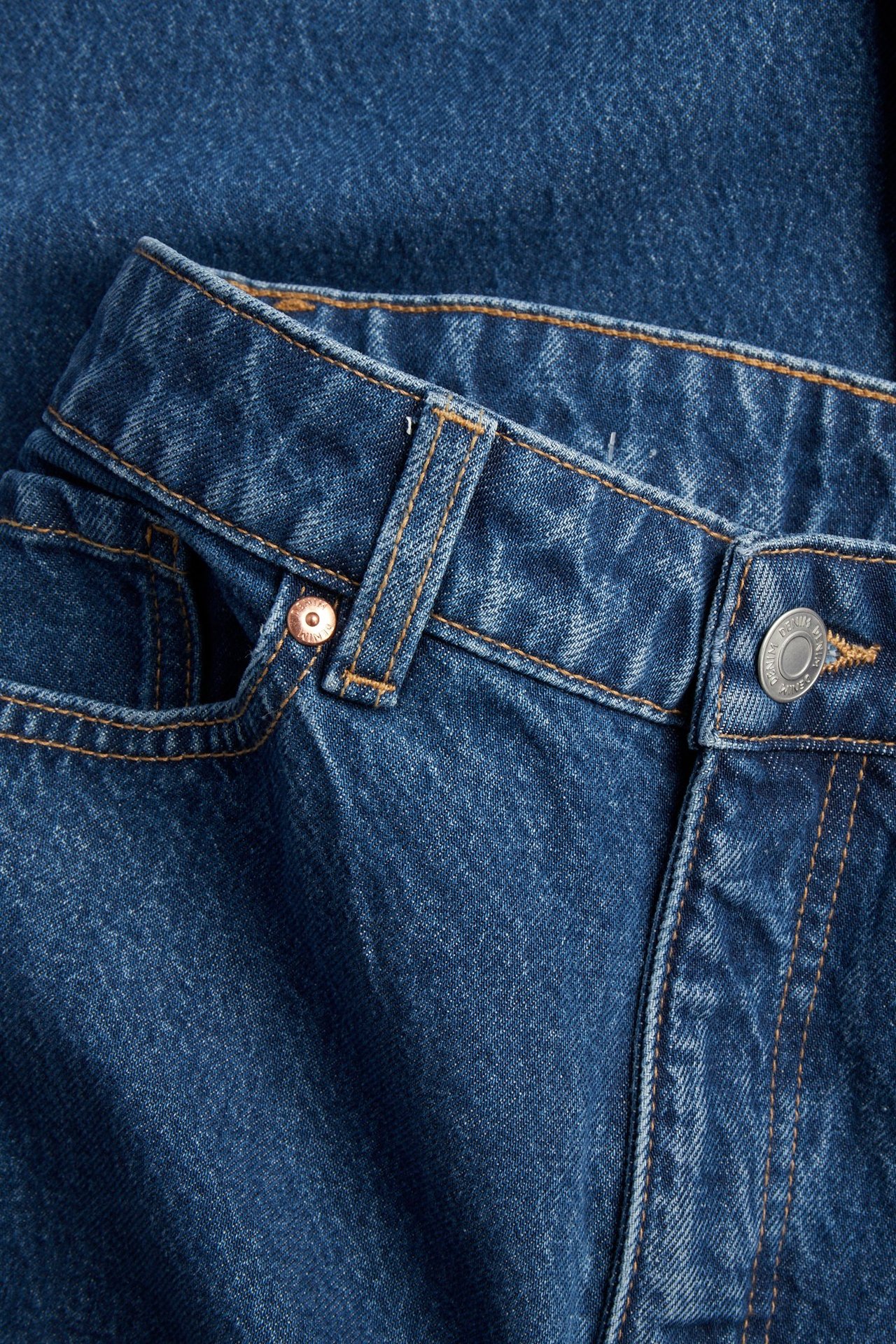 Närbild framifrån på blåa denim high-waist vida jeans med orange sömmar och silverknapp.
