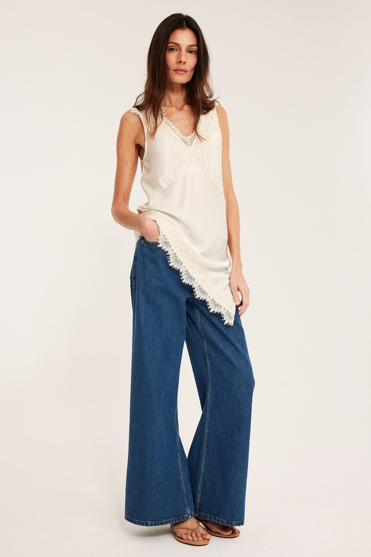 Frontvy av kvinna i vida, högmidjade denimblå jeans, en gräddvit spetslinne och ljusbruna sandaler.