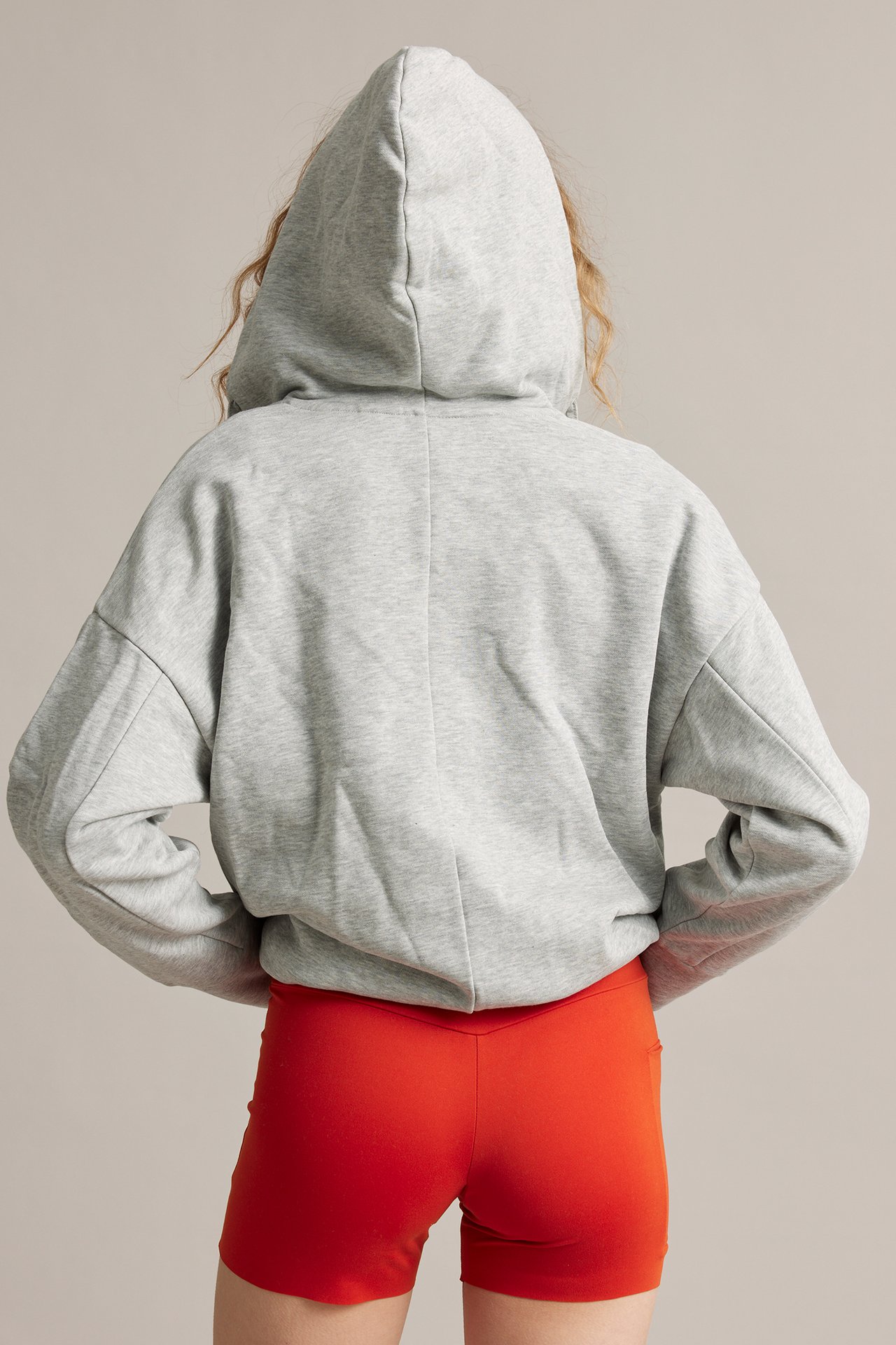 Bakfra av en kvinne i røde, høye bikershorts med sidelomme og en løs grå kort hettegenser.