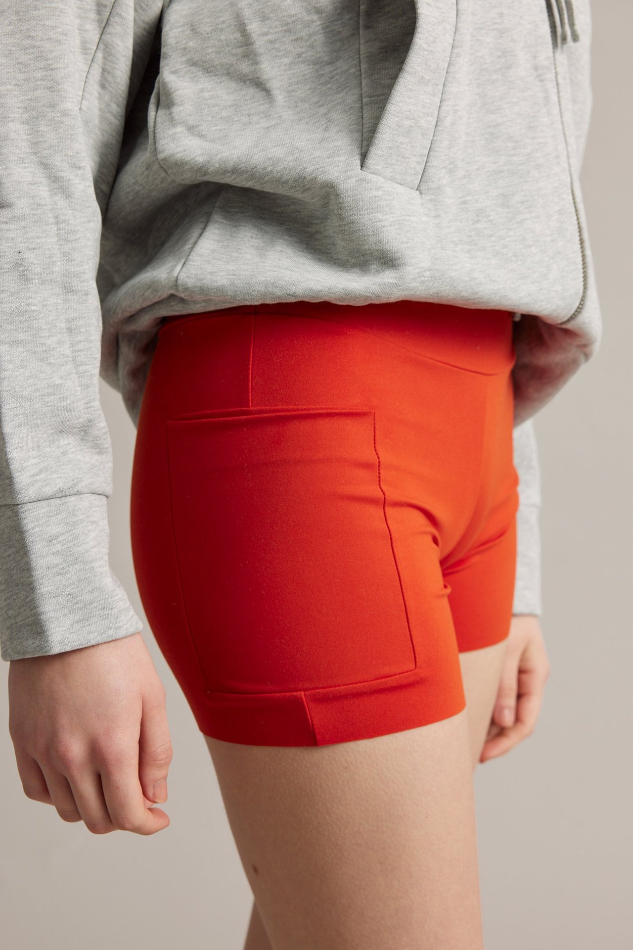 Sidebilde av kvinne i røde, høye stretch-bikershorts med mobillomme og en grå hettegenser.