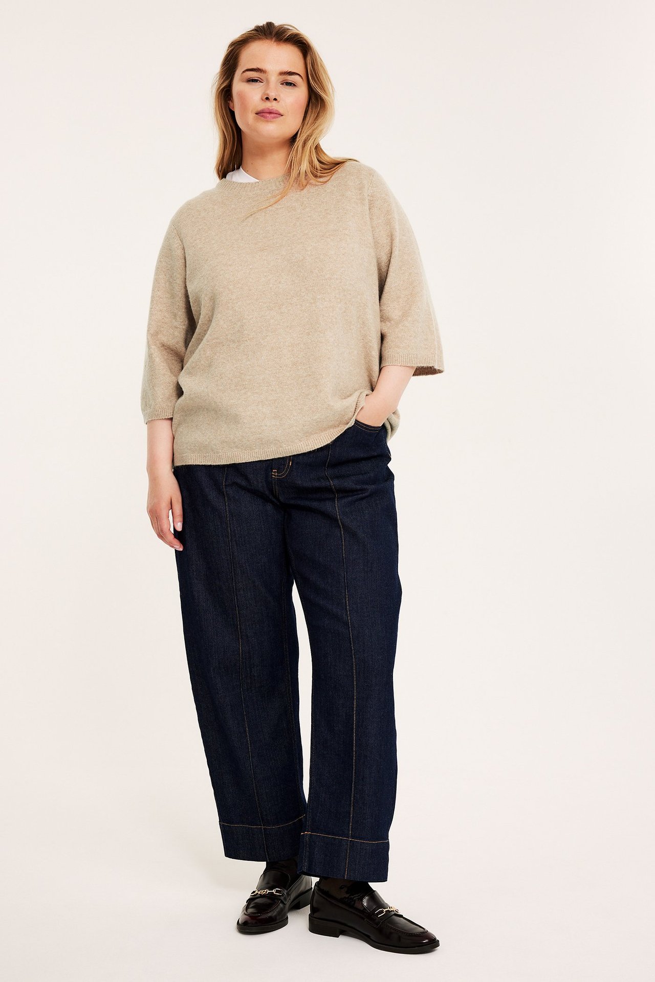 Frontvy av kvinna i beige melerad stickad t-shirt med 3/4-ärm, mörkblå vida jeans och mörka loafers.