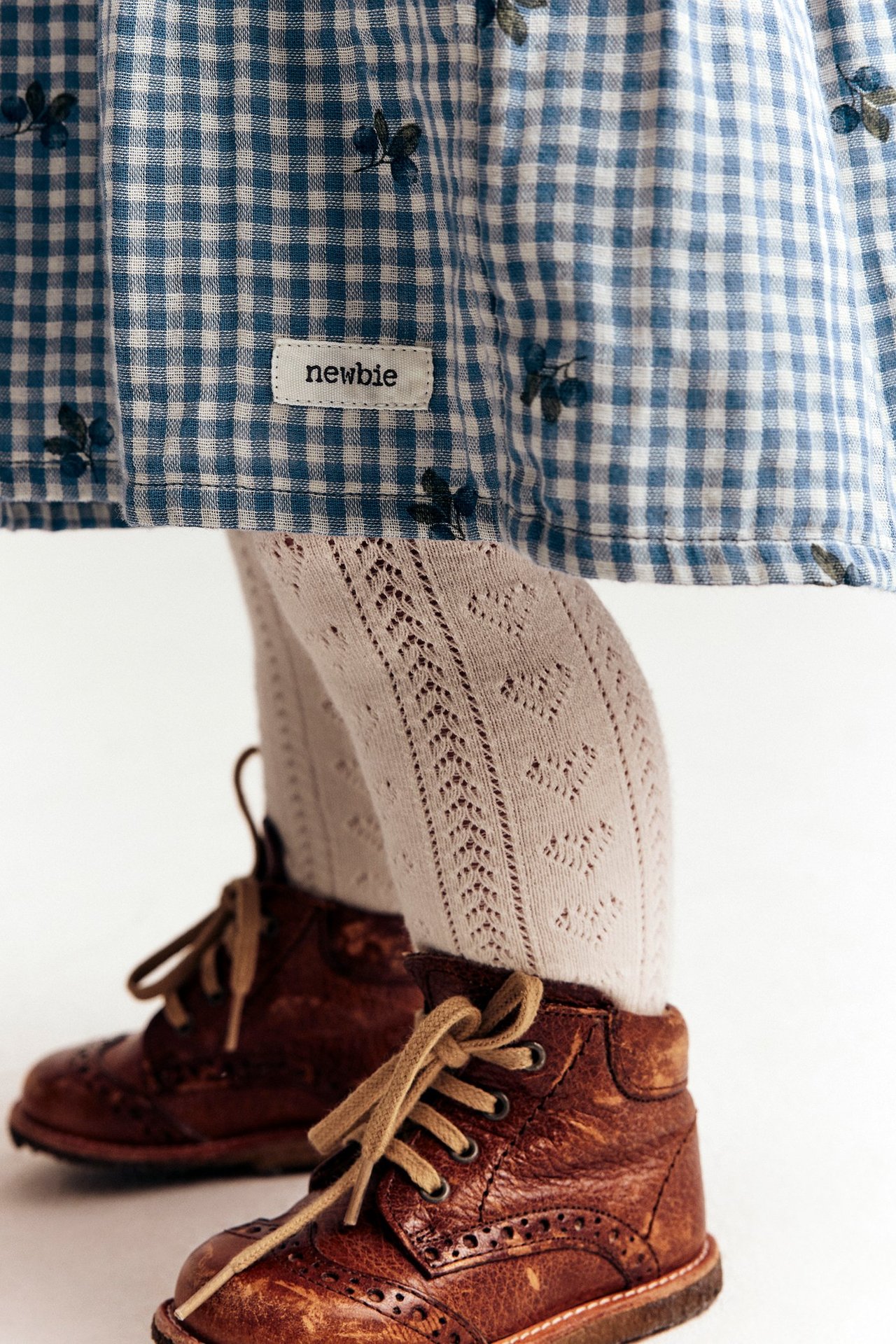 Nærbilde av et barn i blårutet bomullskjole med blåbærtrykk, lyse mønstrede strømpebukser og brune snørestøvler.