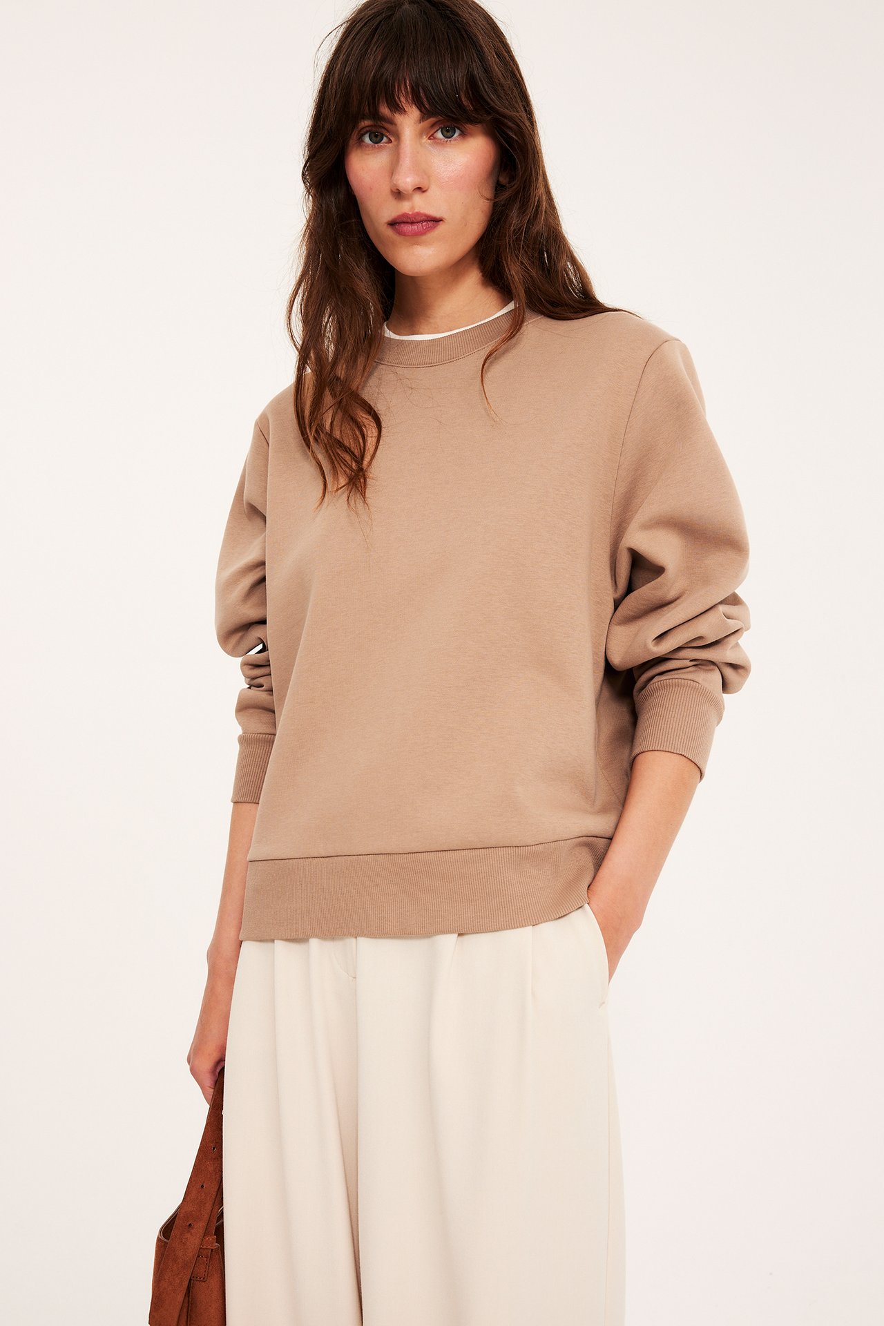 Forfra: kvinne i beige sweatshirt over hvit t-skjorte, kremfargede vide bukser, holder brun veske.