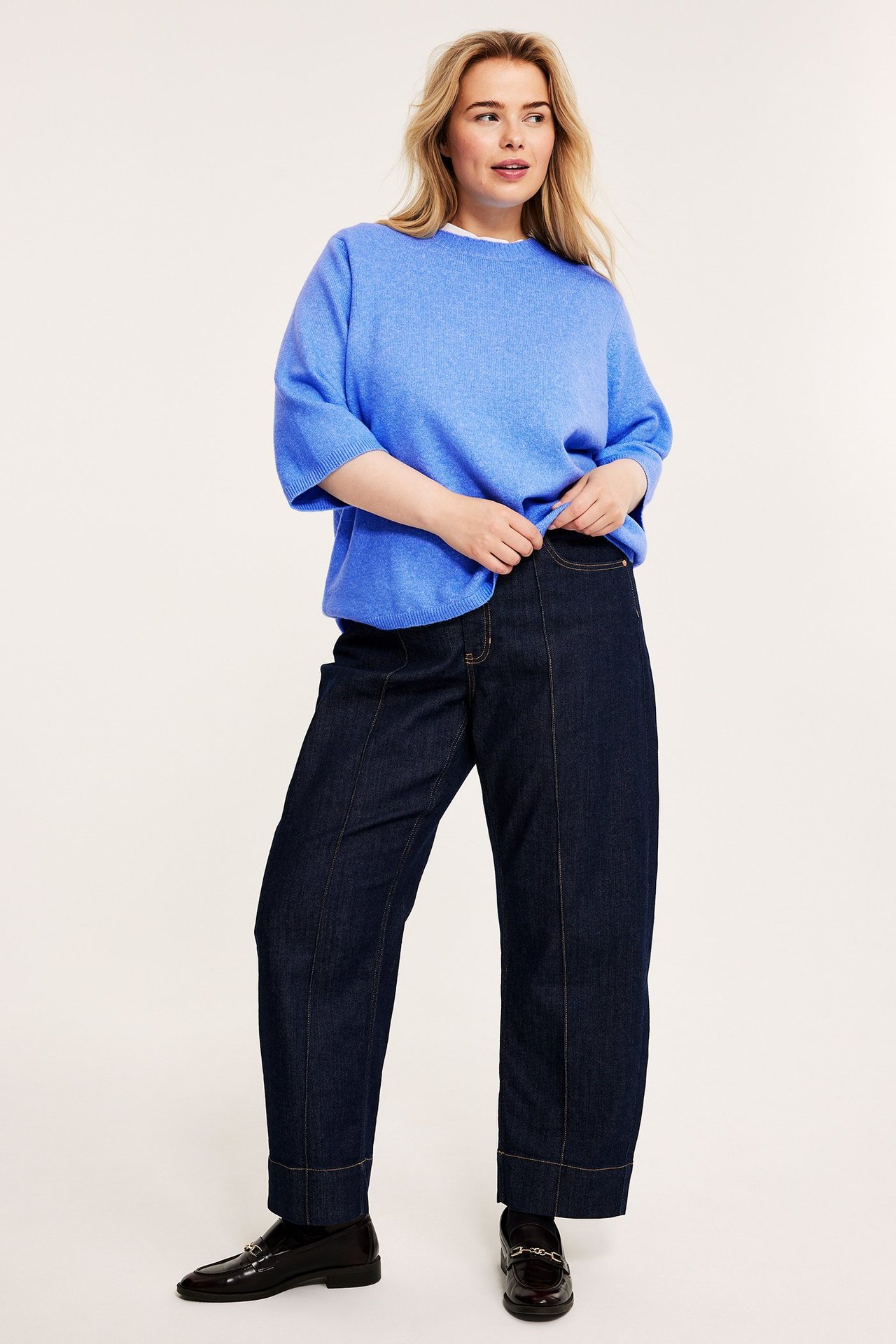 Edestä: Nainen sinisessä 3/4-hihaisessa neulepaidassa, tummansinisissä farkuissa ja tummissa loaferissa.