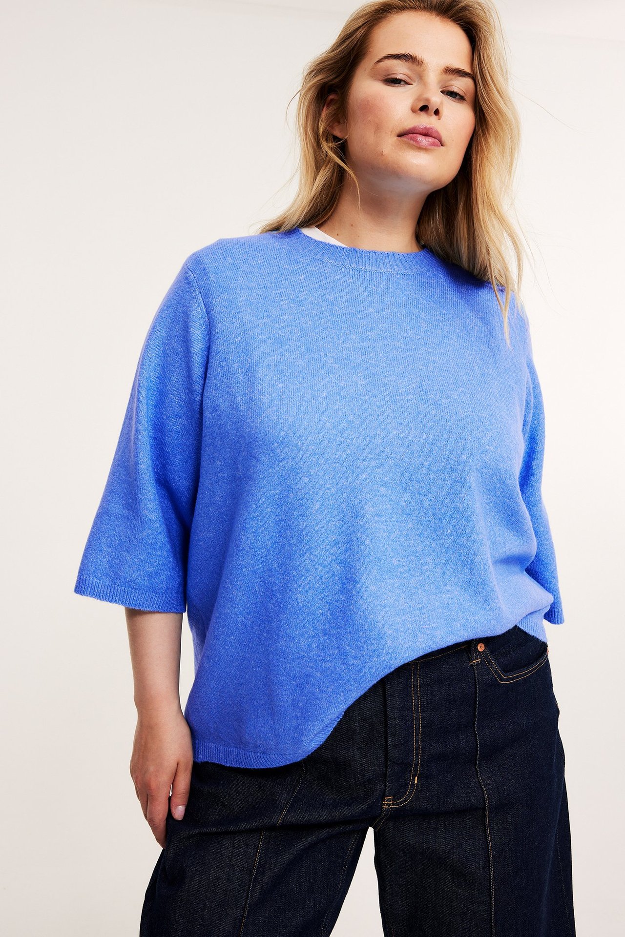 Z przodu: Kobieta w jasnoniebieskim melanżowym dzianinowym t-shircie 3/4 i ciemnych jeansach.