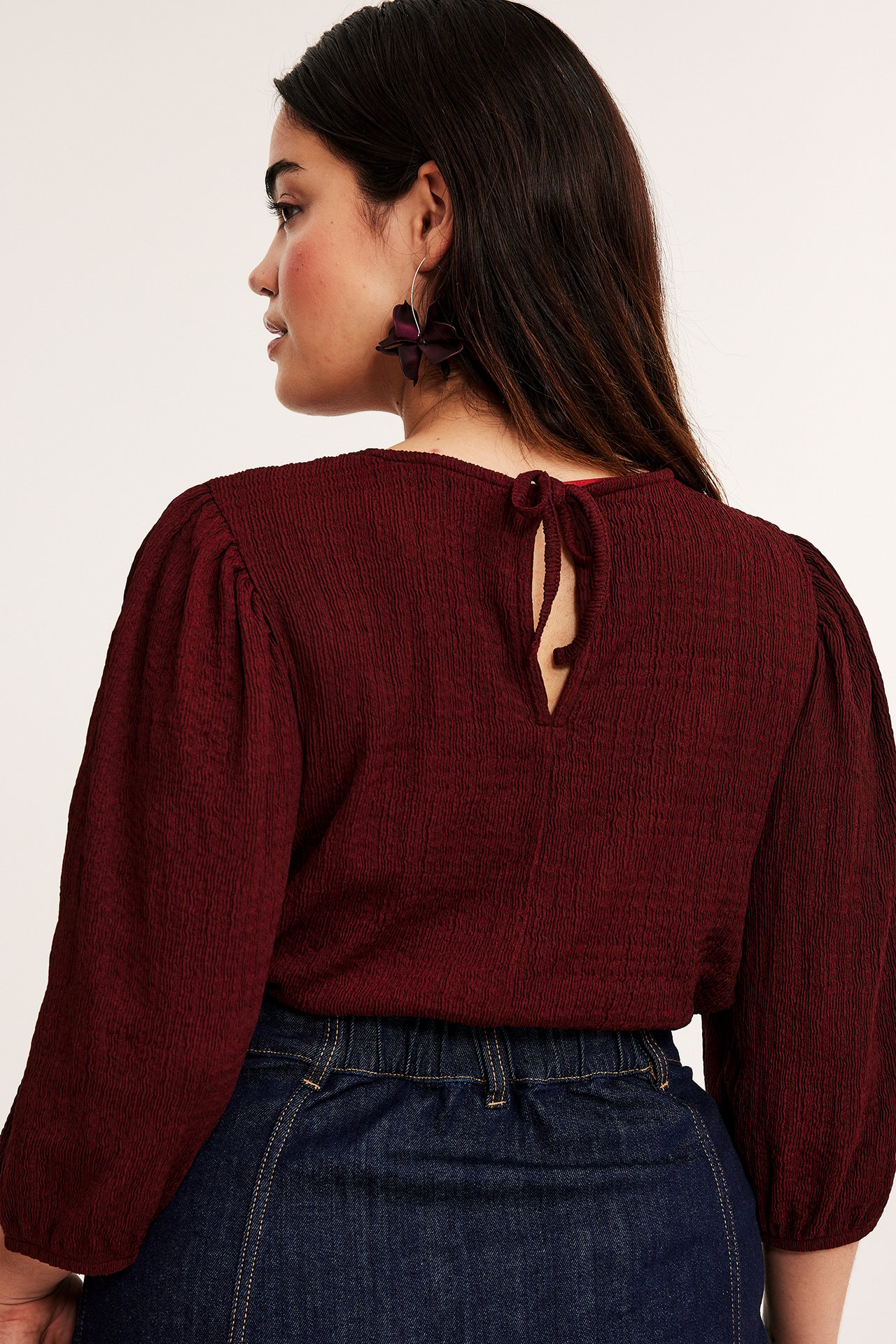 Bakifrån: kvinna i mörkröd strukturerad topp med puffärm, knytband och mörkblå jeans.