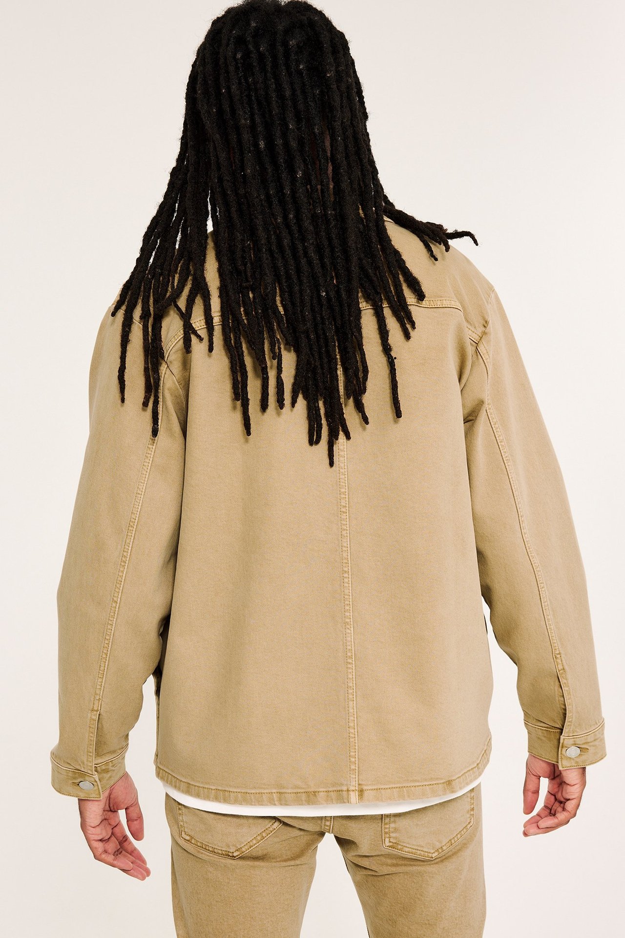 Widok z tyłu: mężczyzna w beżowej kurtce z twillu bawełnianego, białym podkoszulku i beżowych spodniach.