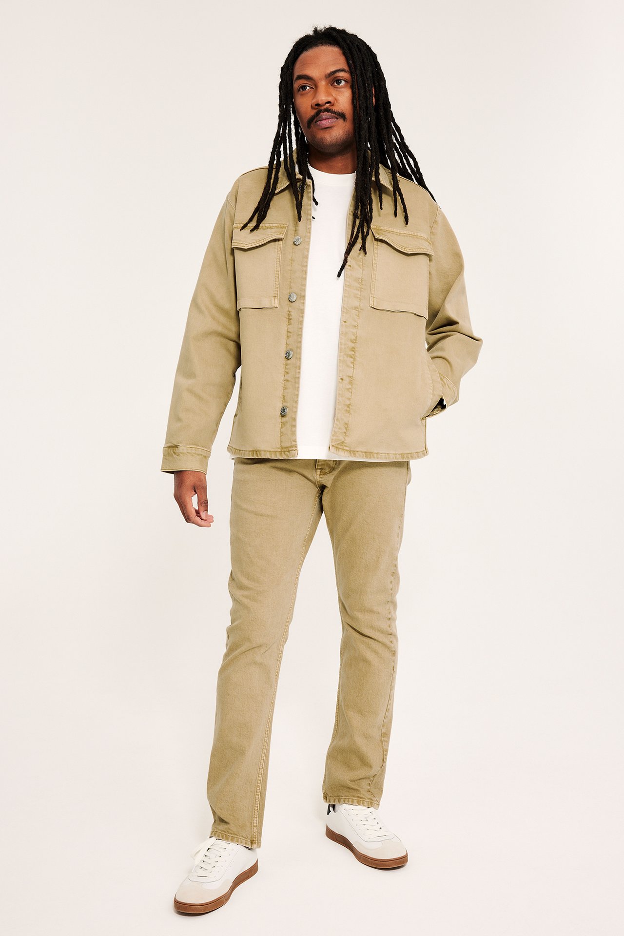 Edestäpäin: mies beigessä puuvillatwillitakissa isoilla taskuilla, valkoinen t-paita, beiget farkut, valkoiset tennarit.