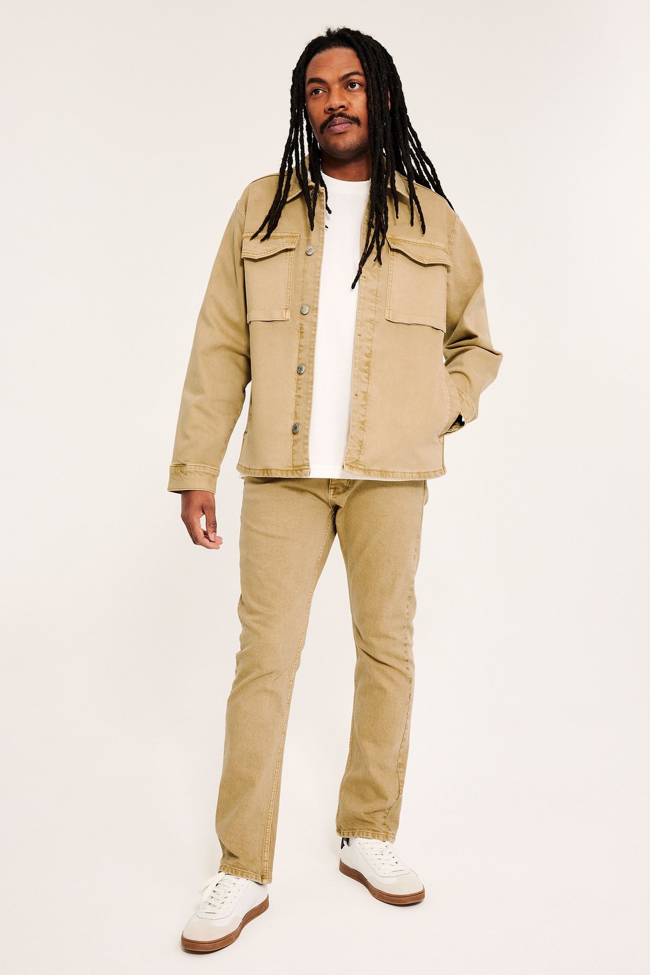 Frontvy: man i beige bomullstwilljacka med fickor, vit t-shirt, beiga byxor och vita sneakers.