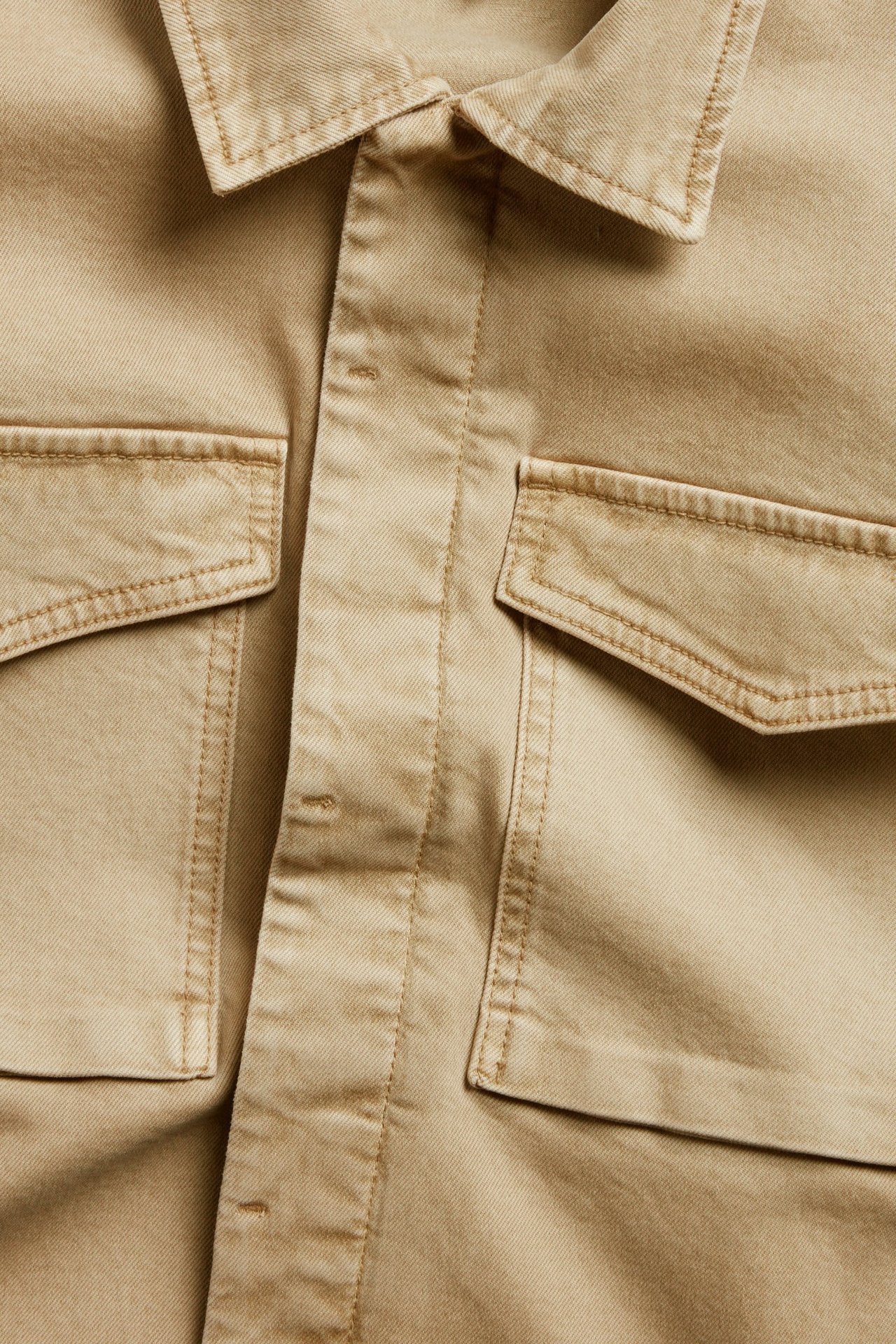 Zbliżenie beżowej kurtki z diagonalu bawełnianego z kołnierzem, zapięciem na guziki i dwiema kieszeniami.