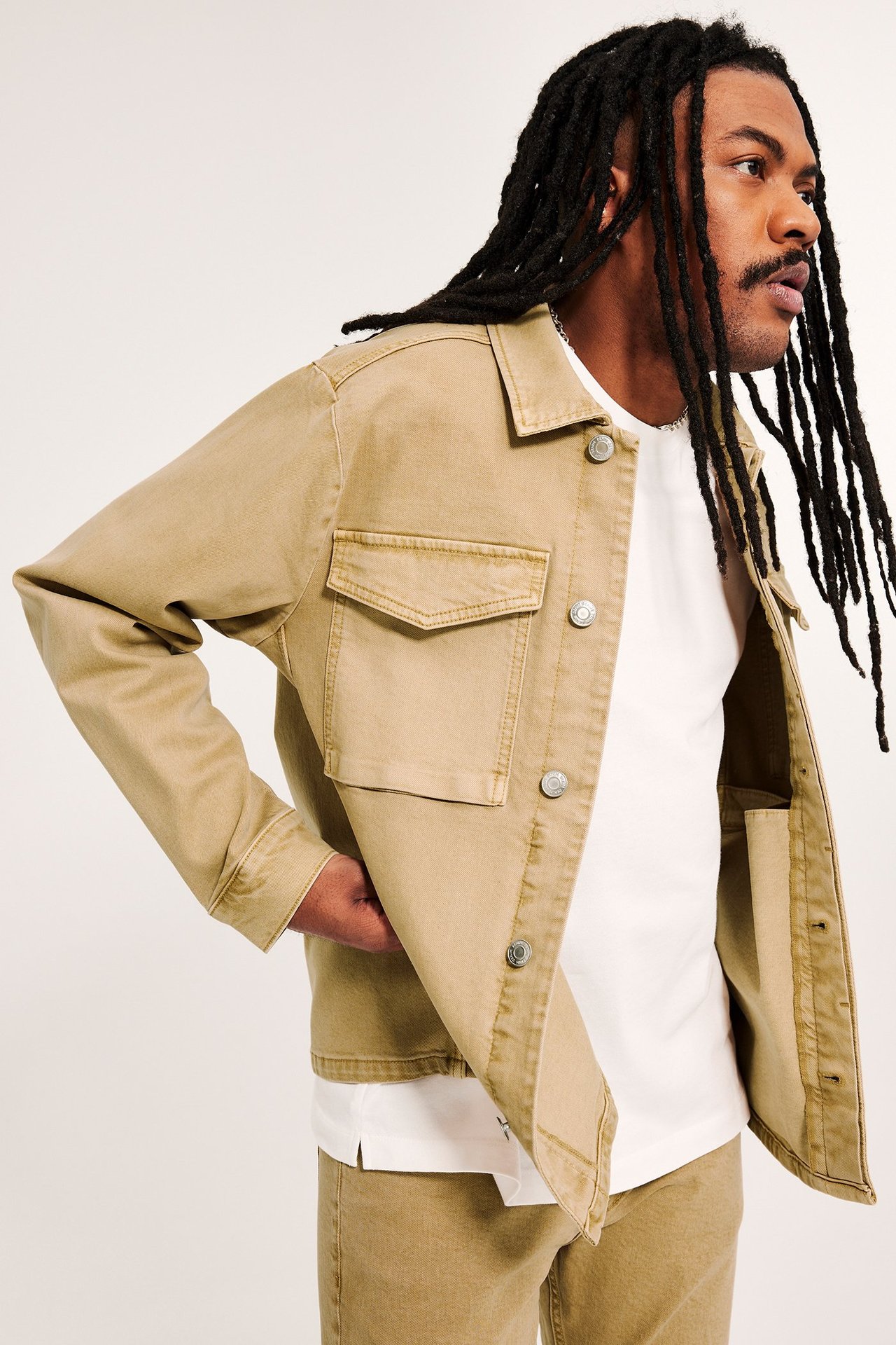 Sidovy: man i öppen beige bomullstwilljacka med stora fickor, vit t-shirt och beiga byxor.
