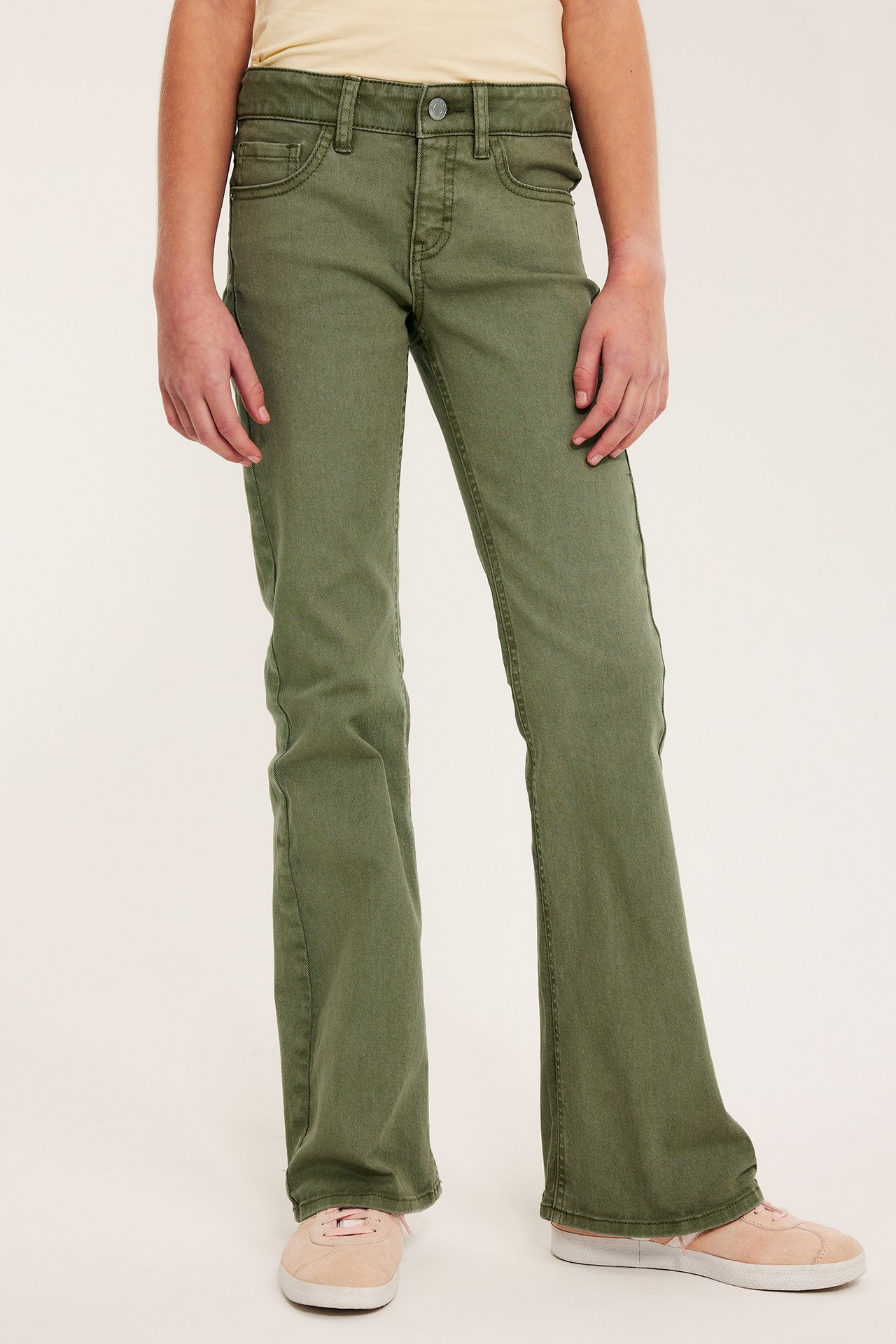 Forfra: et barn i mørkegrønne bootcut-jeans med knappelukking, lys gul topp og rosa joggesko.